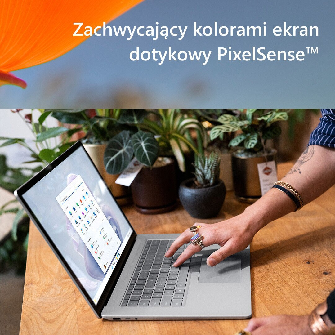 Osoba korzystająca z laptopa na drewnianym stole. Widoczne są rośliny i tekst.