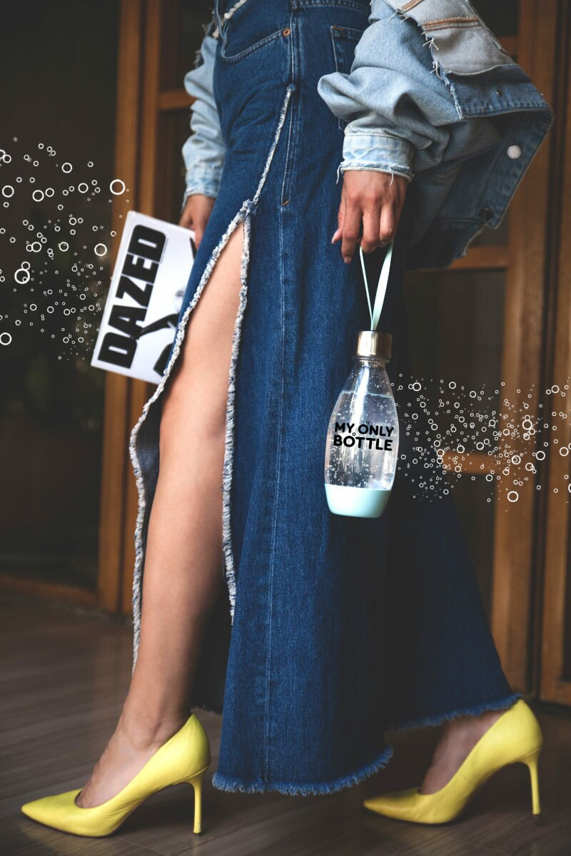 Osoba w dżinsach trzymająca butelkę wody i magazyn, w żółtych obcasach.