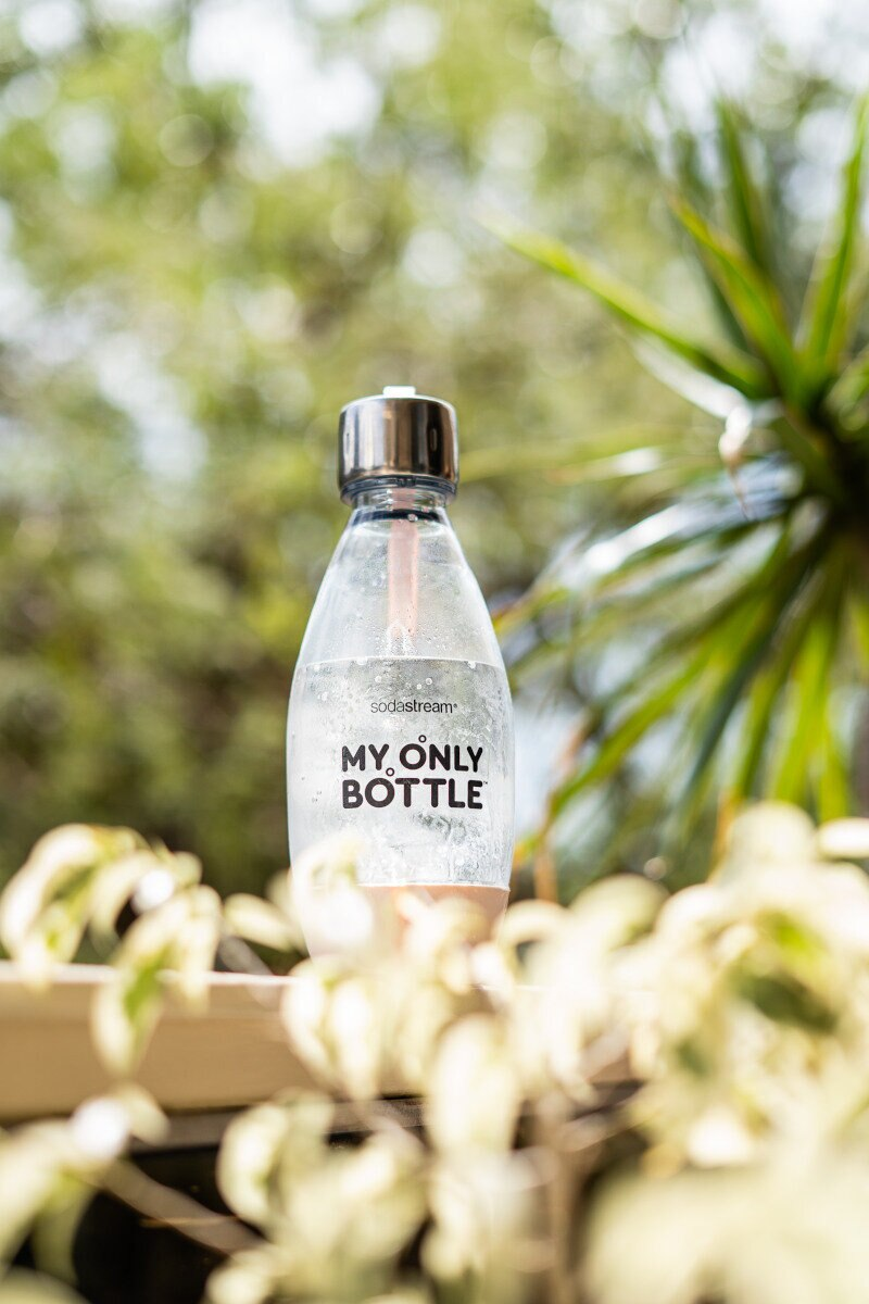 Butelka SodaStream ze srebrną nakrętką i różową słomką, na tle zielonych roślin i liści.