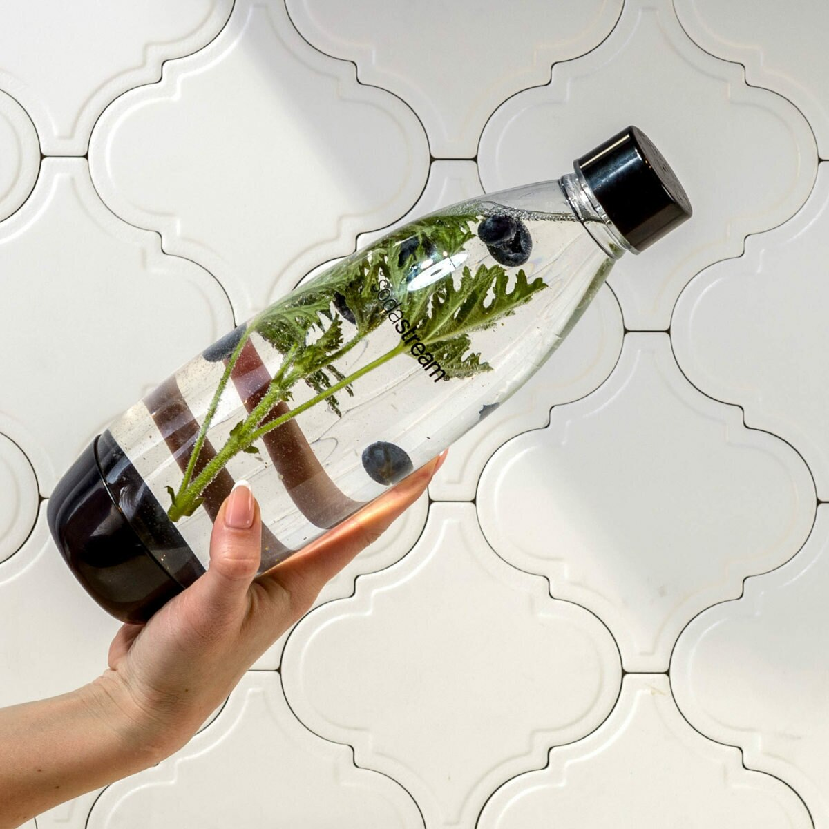 Przezroczysta butelka z wodą, jagodami i zielonymi liśćmi, trzymana przez dłoń na tle ściany z płytek.