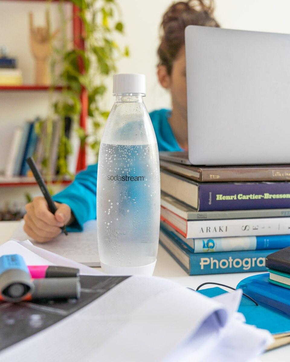Osoba pisząca obok butelki SodaStream, książek i papierów. Biały stół, rozmyte tło.
