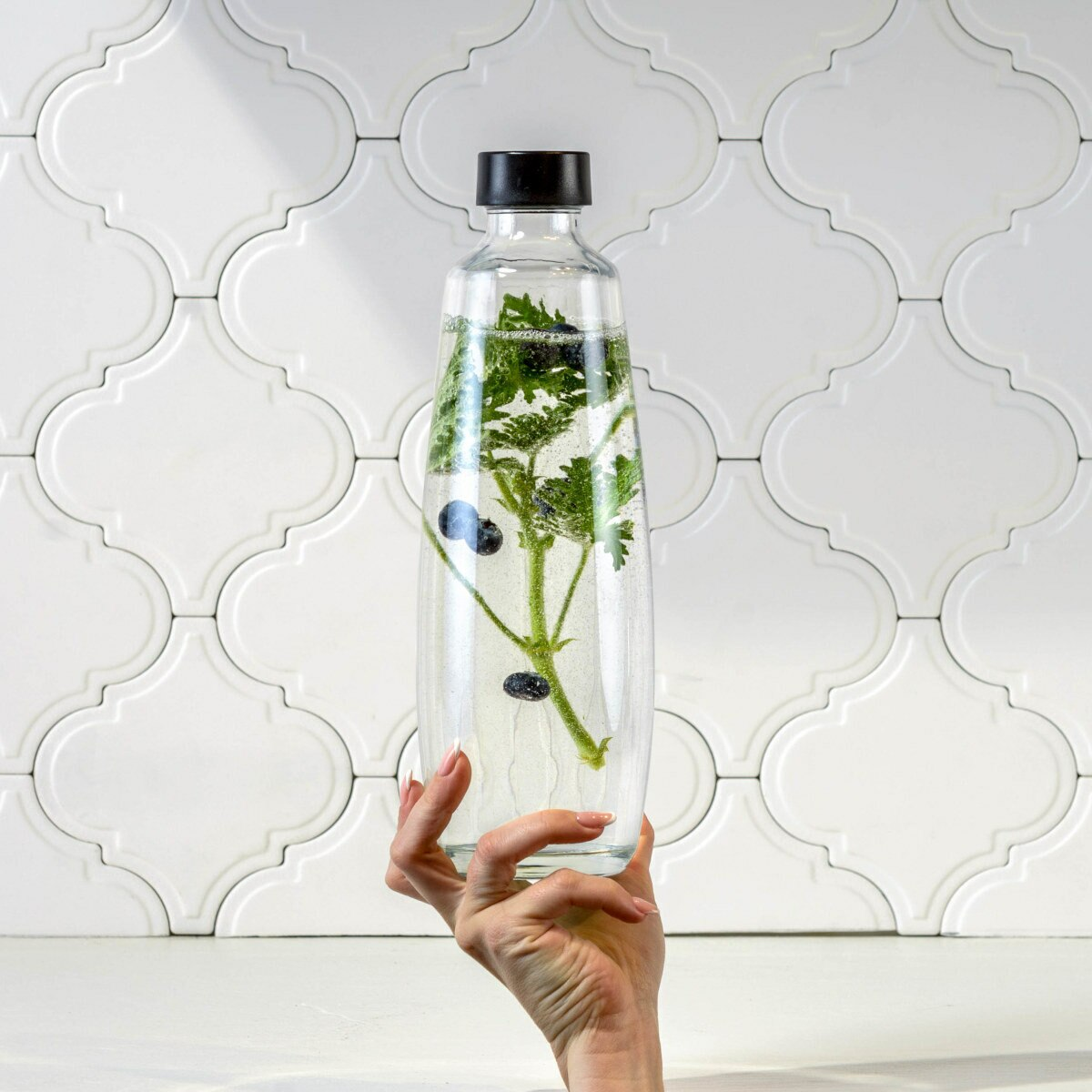 Ręka trzymająca przezroczystą butelkę z roślinami i jagodami na tle białych płytek.