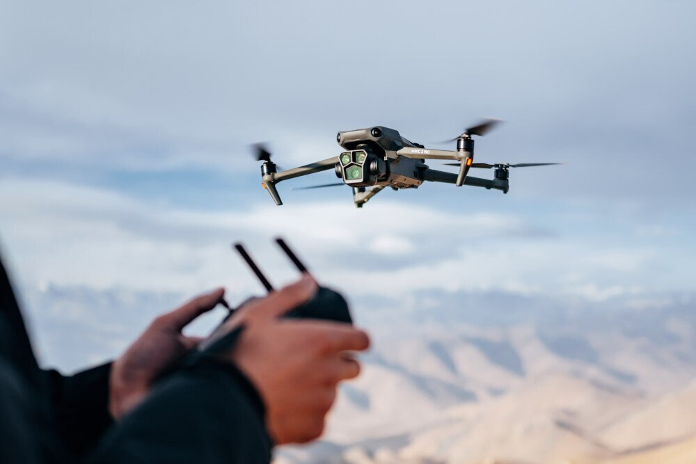 Dron w locie, trzymany przez ręce. Rozmyte tło, błękitne niebo i brązowe wzgórza.