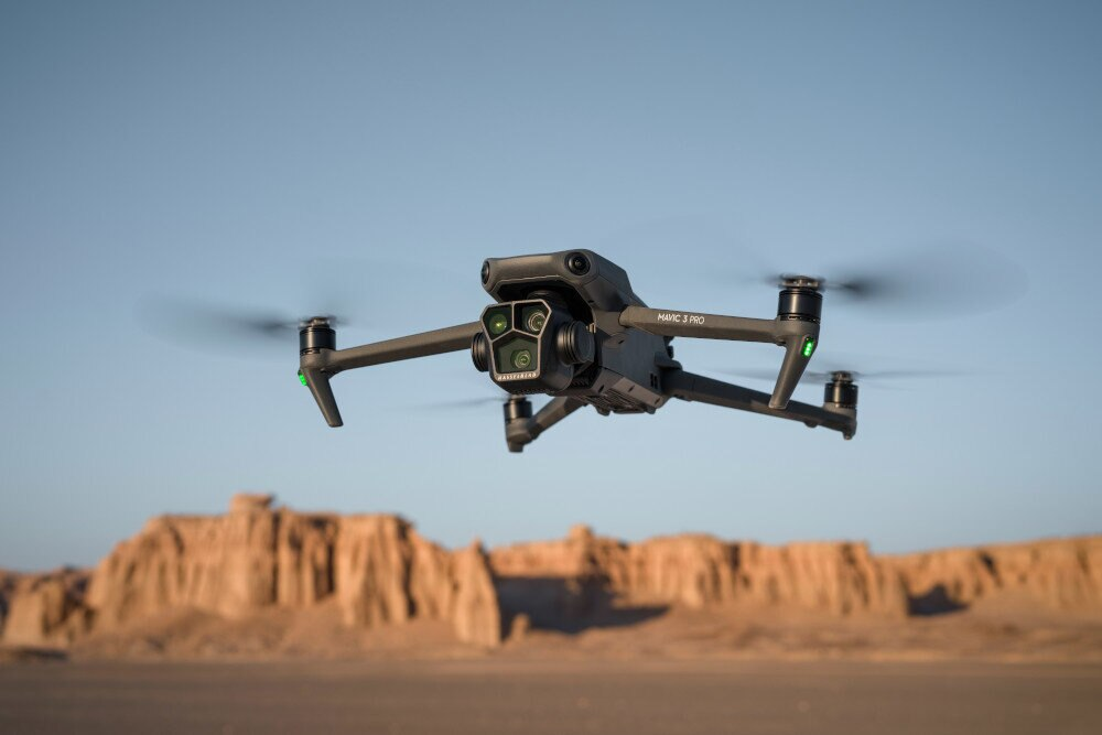 Dron lecący nad pustynnym krajobrazem. Dron jest szary i skierowany w stronę kamery. Niebo jest niebieskie.