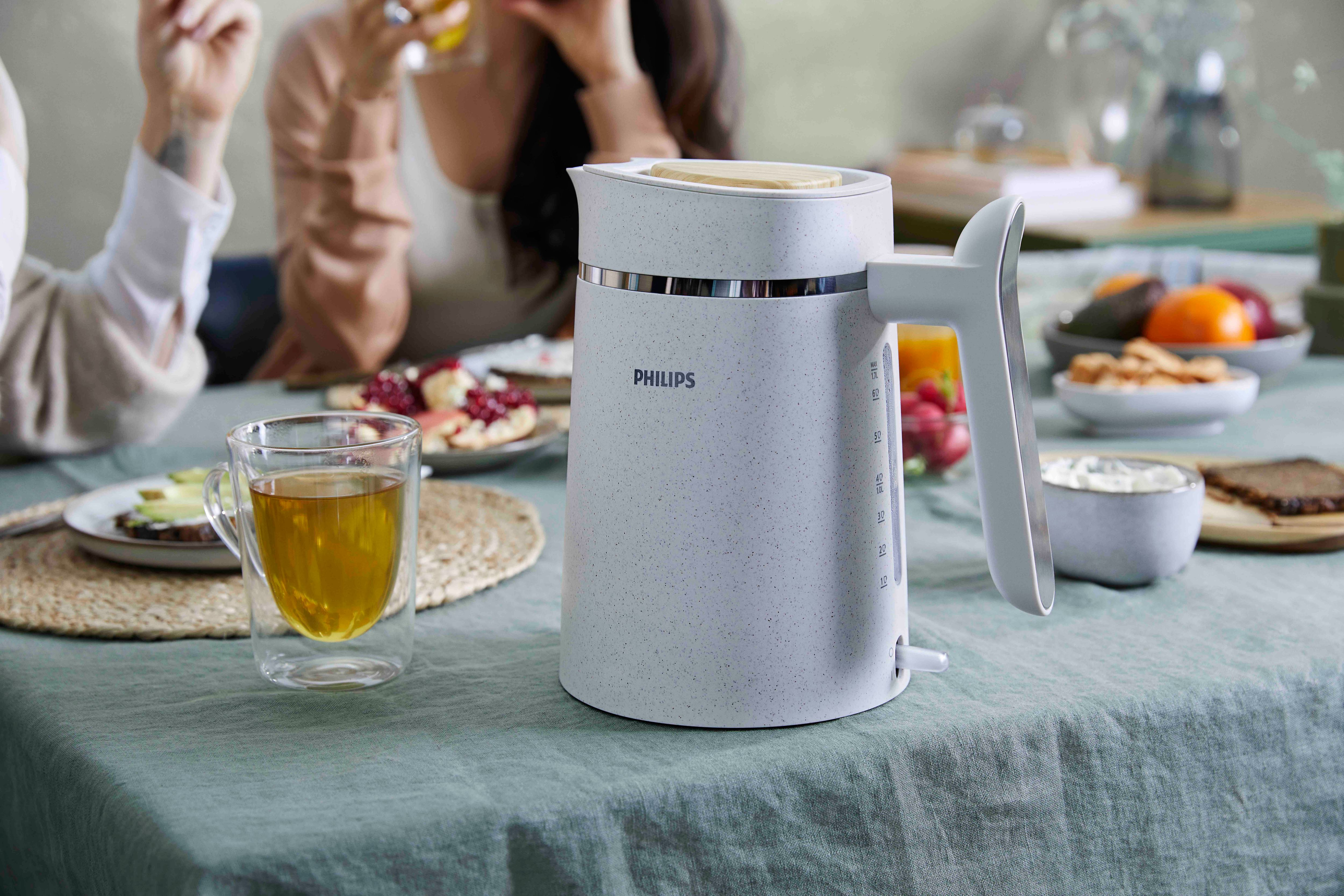 A white and gray Philips kettle on a table, with people and food in the background.