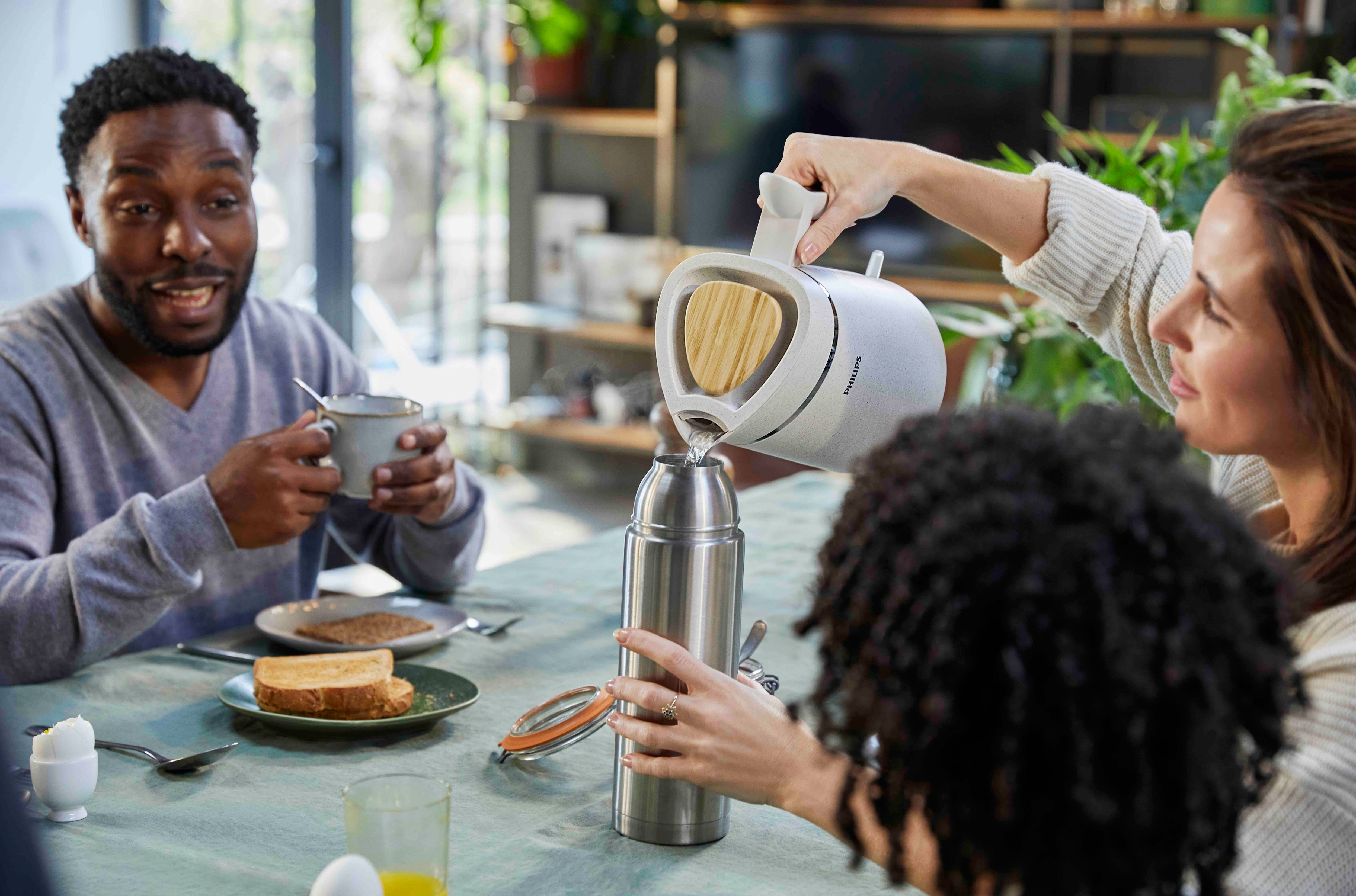 People at a table. One is pouring water from a white kettle into a silver thermos, with toast and eggs.