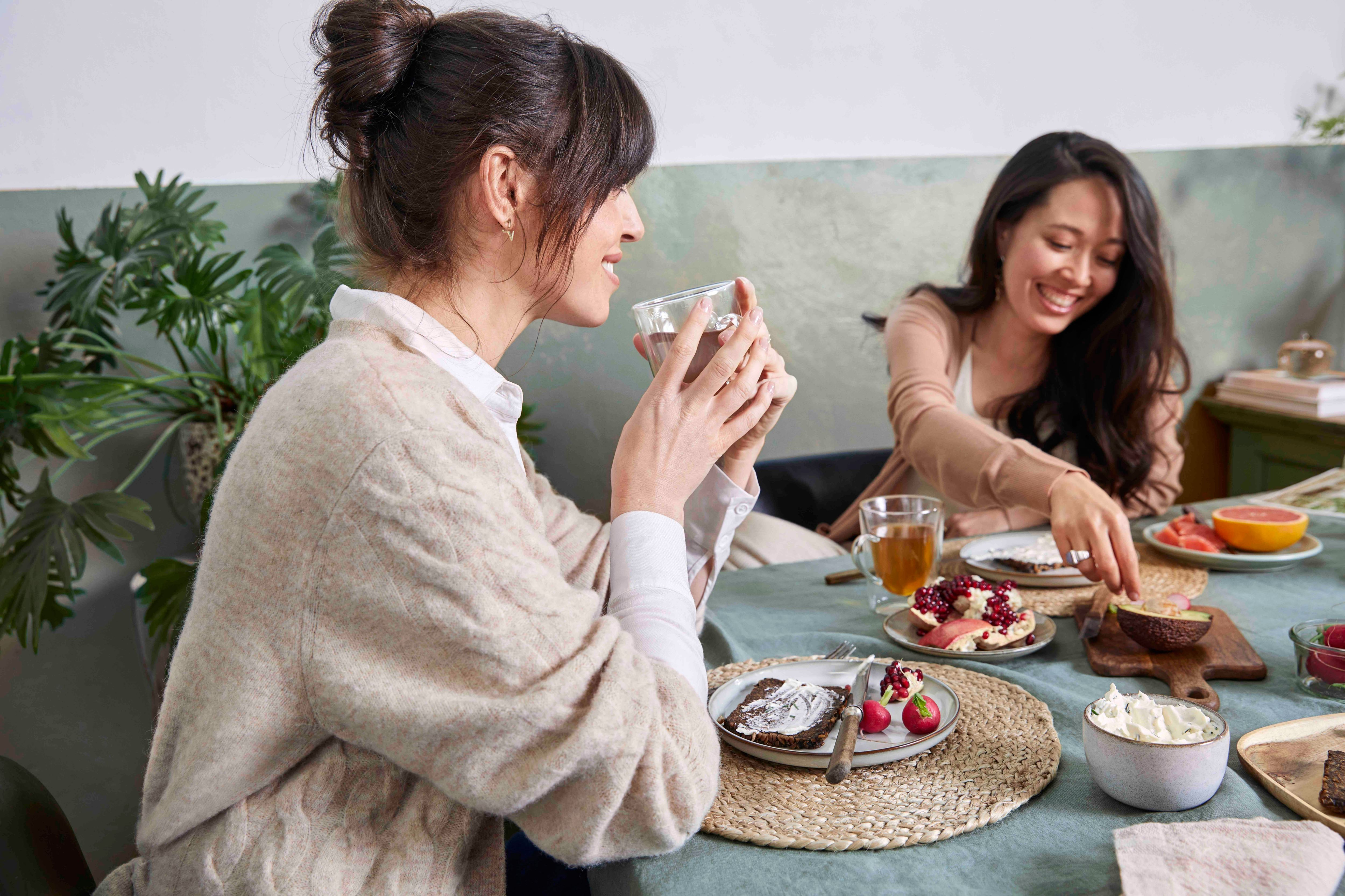 Two women at a table, one drinking, the other eating. Table has food, drinks, and a floral arrangement.