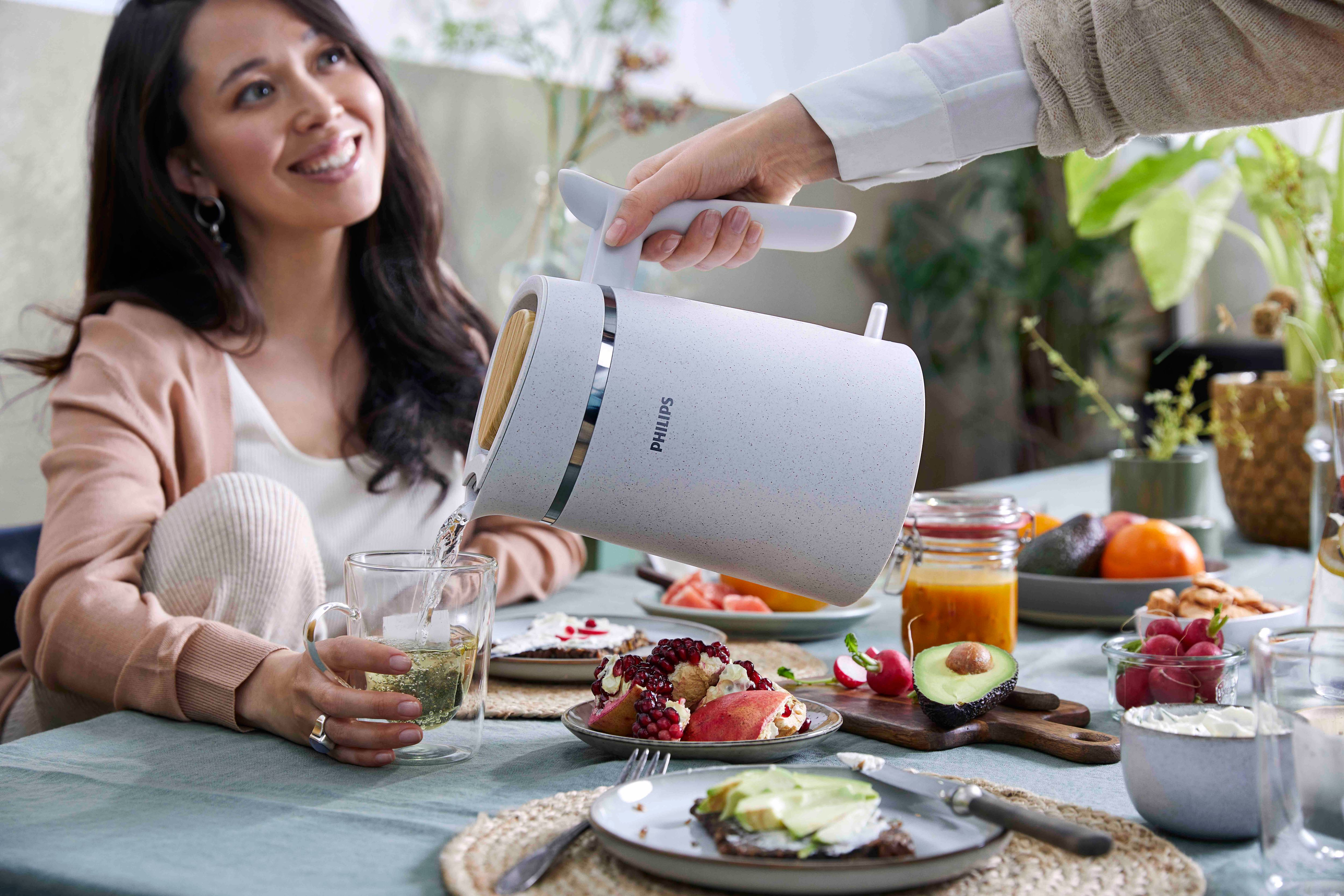 A woman pours water from a Philips kettle into a glass. Food and plants are on the table.