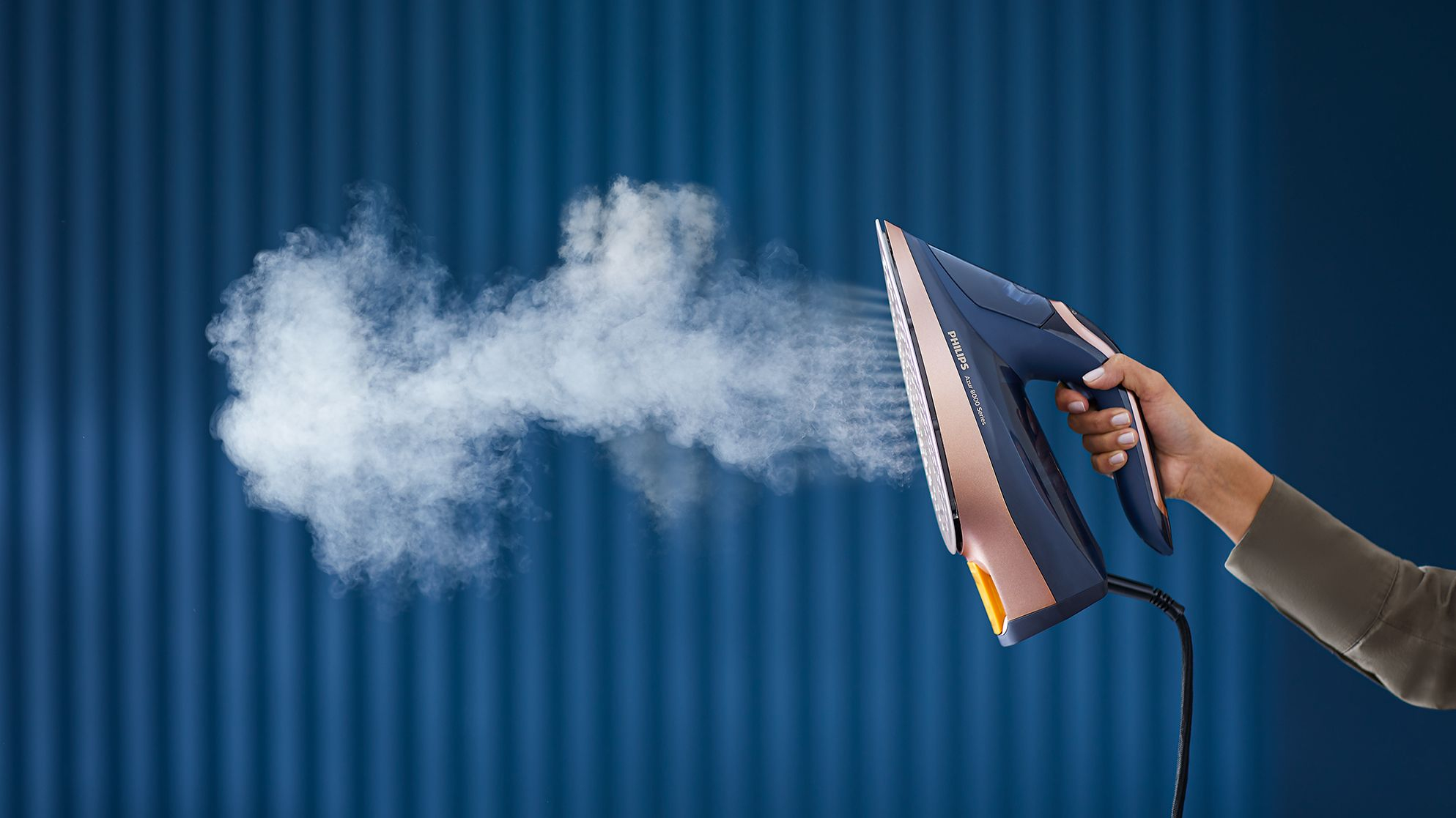 Main d'une personne tenant un fer à repasser bleu et cuivré, de la vapeur sortant.
