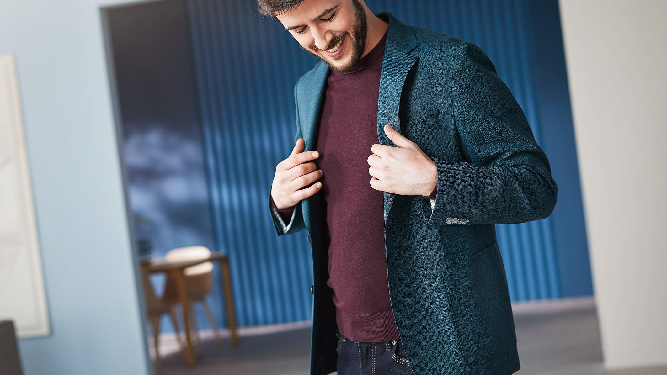 Homme souriant, ajustant une veste bleu-vert sur un pull bordeaux et un jean.