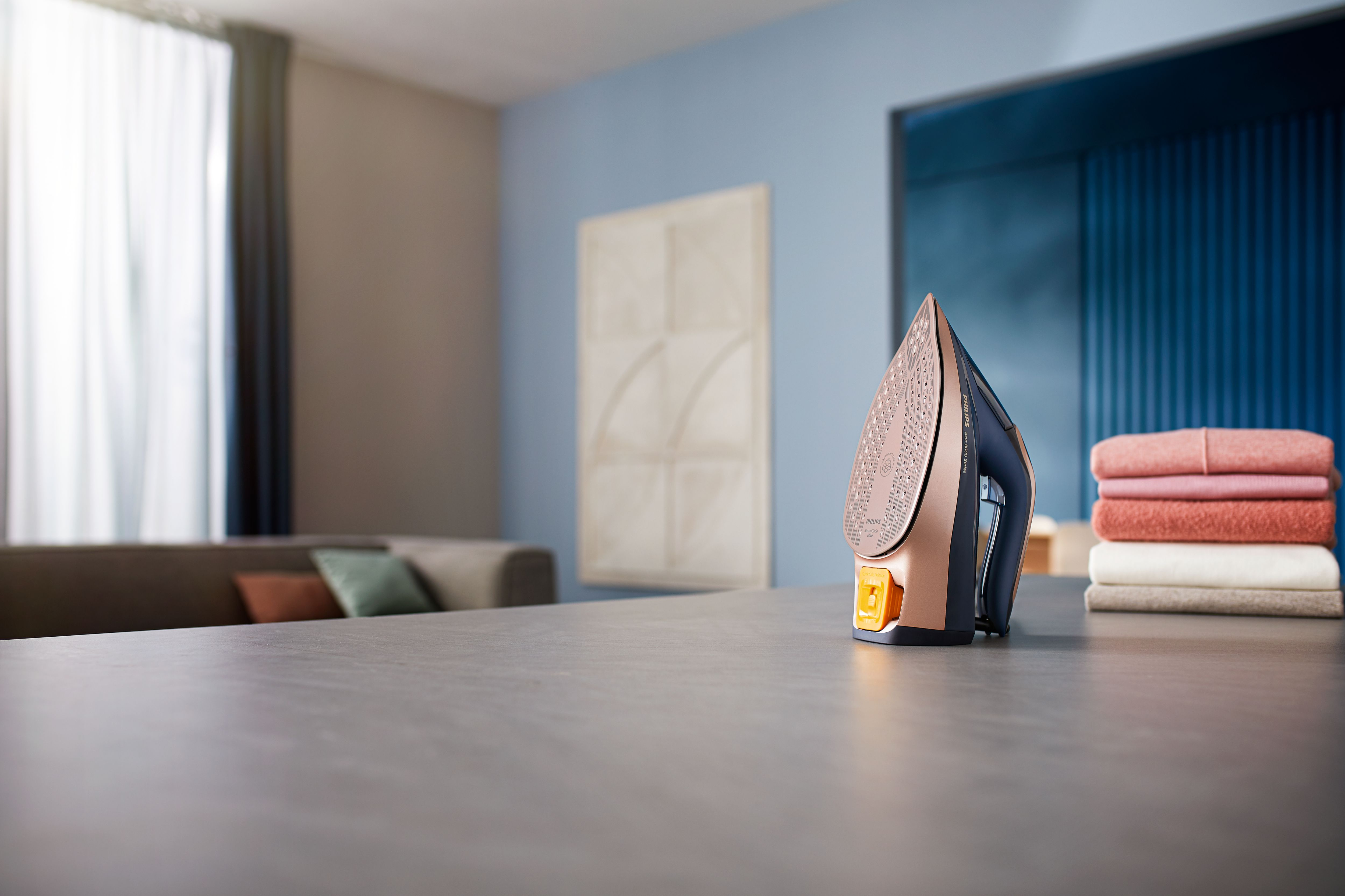 Un fer à repasser sur une table avec des vêtements pliés, un canapé en arrière-plan.