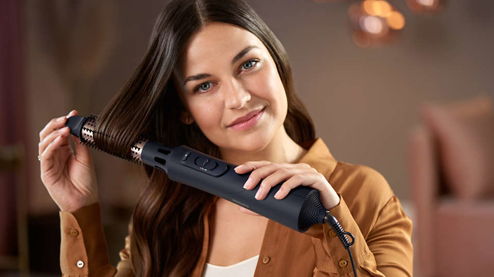 Woman holding a black hair styler, smiling, brown shirt. Soft background.