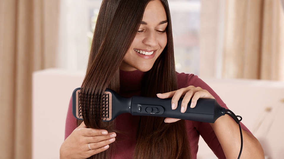 Woman using a hair styling brush on her hair, smiling. Burgundy top.