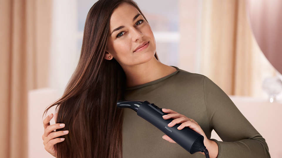 A woman with long hair uses a black hair dryer in front of a window.