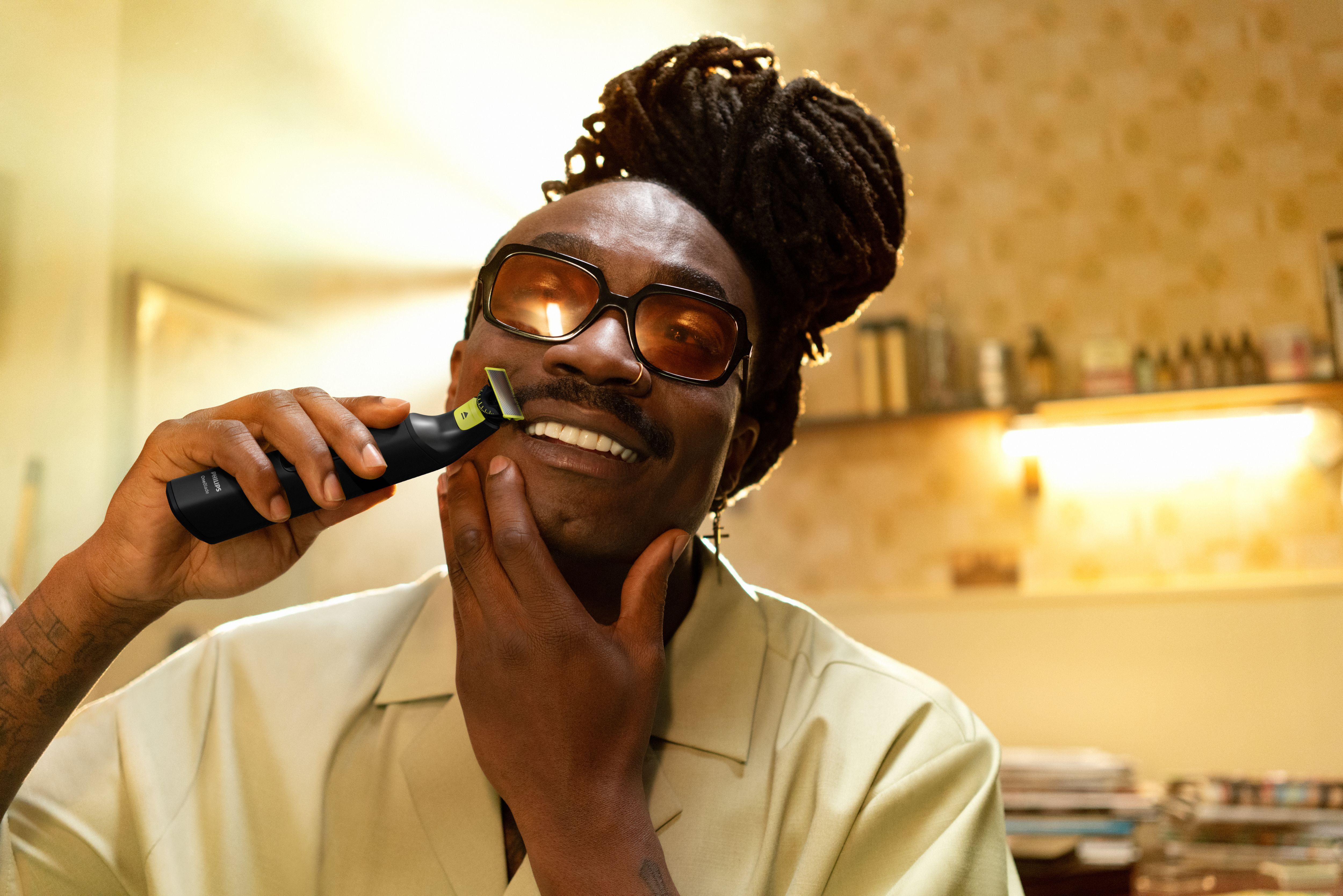 Een man scheert zijn baard met een trimmer. Hij draagt een bril en een beige shirt. De achtergrond is een badkamer.