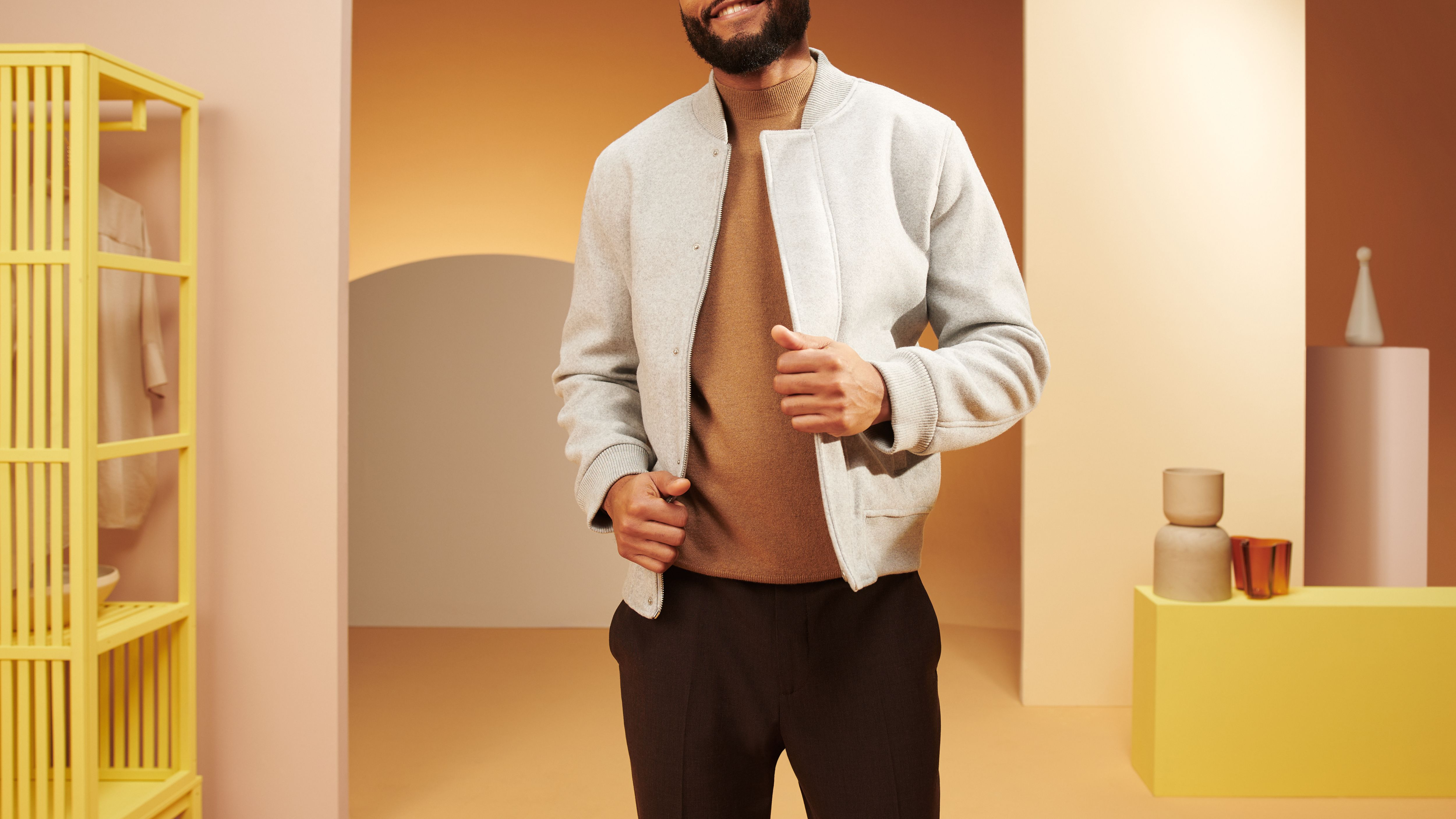 Homme en veste grise et col roulé marron, debout. Étagère jaune et décor en arrière-plan.