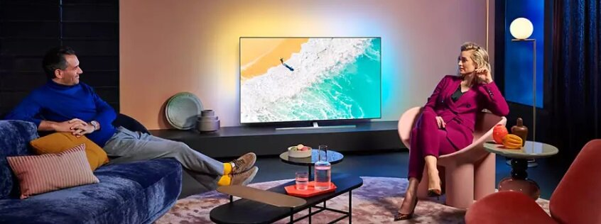 Salon z telewizorem pokazującym scenę na plaży. Mężczyzna i kobieta siedzą i patrzą na telewizor.