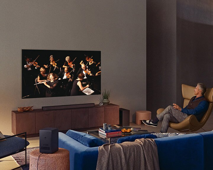 Mężczyzna ogląda telewizor z orkiestrą. W pokoju soundbar, subwoofer i niebieska sofa.