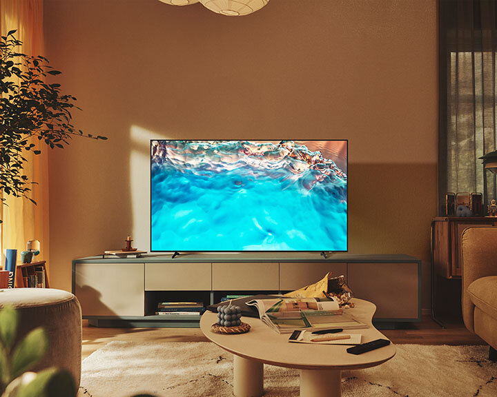 Salon z telewizorem wyświetlającym scenę błękitnego oceanu. Stolik kawowy z czasopismami. Beżowa sofa i pufa. Ciepłe oświetlenie.