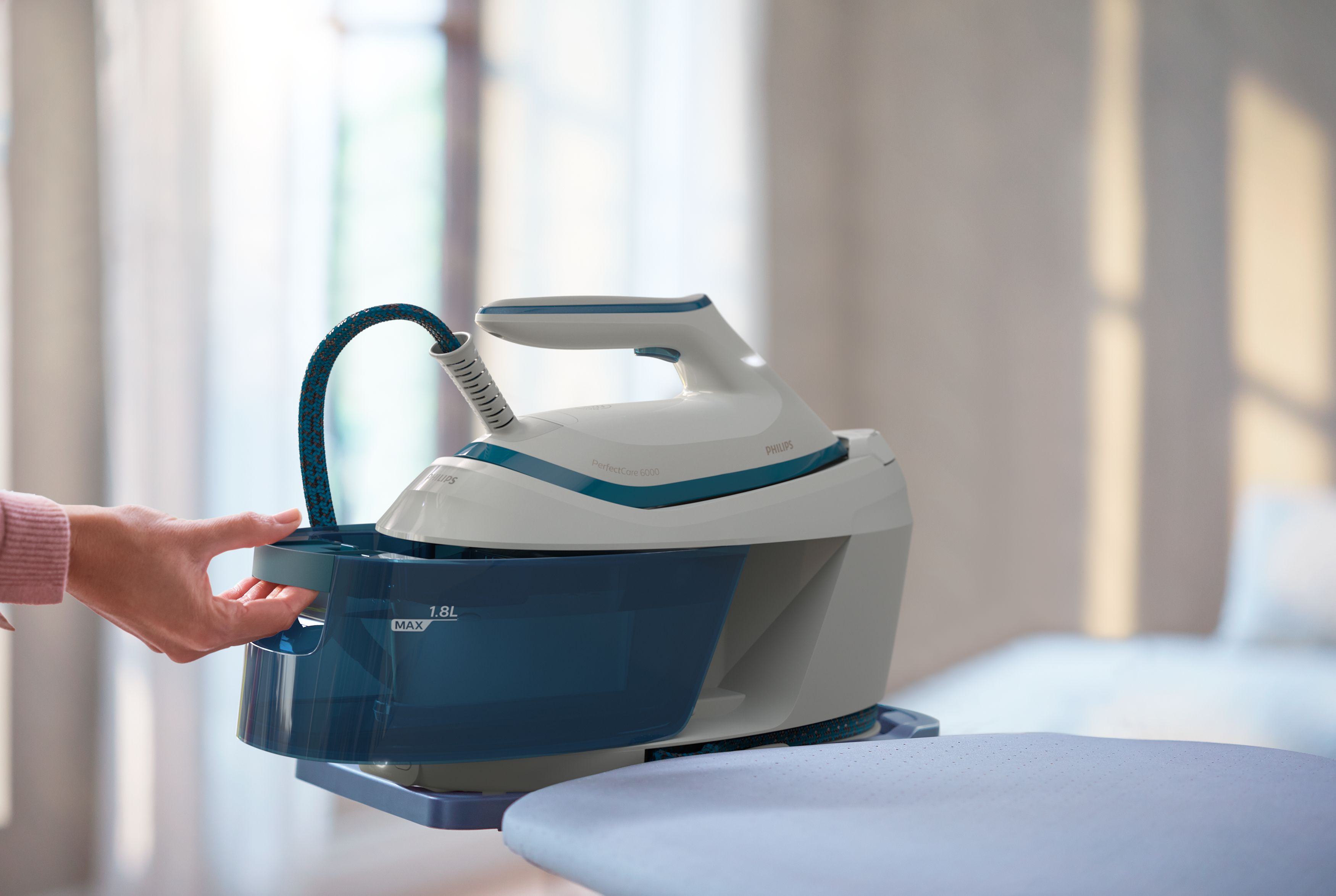 A white and blue steam iron with a water tank on an ironing board. A hand is filling the tank.