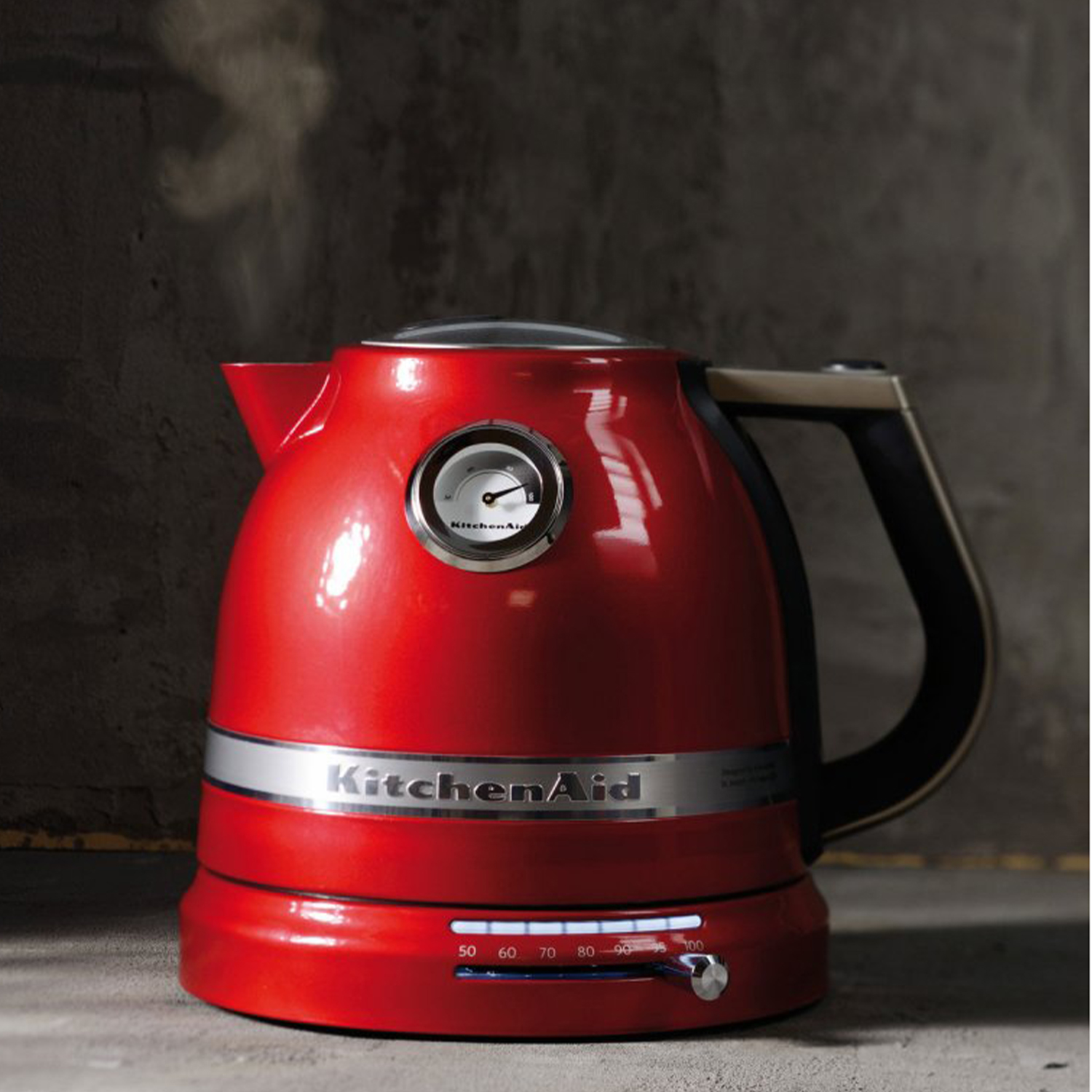 Un bollitore KitchenAid rosso con un indicatore di temperatura. Vapore esce dal beccuccio. È su un bancone di pietra.
