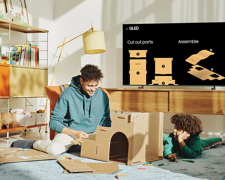 Dwóch chłopców buduje kartonowy zamek. Jeden siedzi, drugi leży. Telewizor wyświetla schematy zamku.