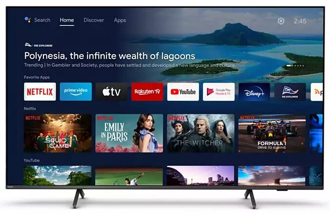 Duży telewizor wyświetlający ekran główny z ikonami aplikacji. Telewizor stoi na stojaku w salonie.