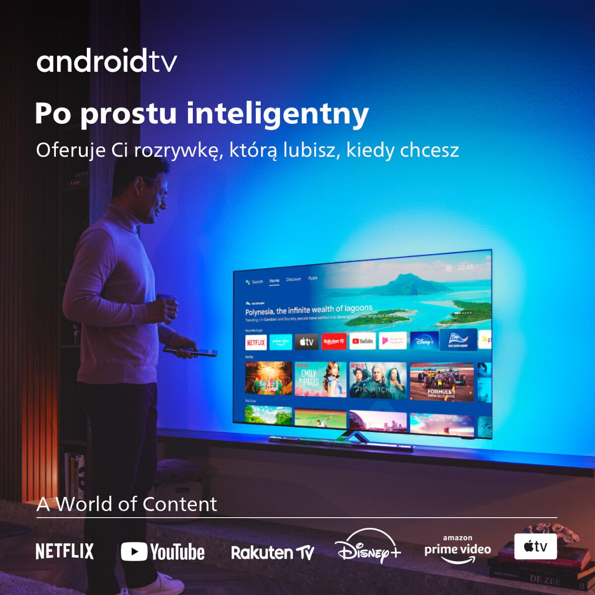 Mężczyzna stoi przed dużym telewizorem z aplikacjami. Widoczne logo Android TV. Na ekranie wyświetlane są aplikacje.