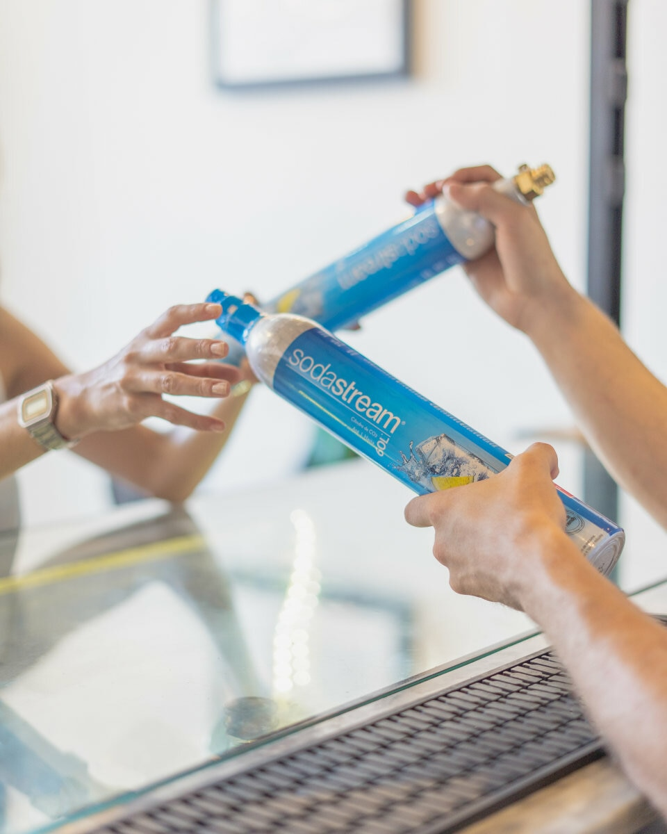 Dłonie podają niebieskie butelki Sodastream. Jedna ręka ma zegarek. Szklana powierzchnia i biała ściana w tle.