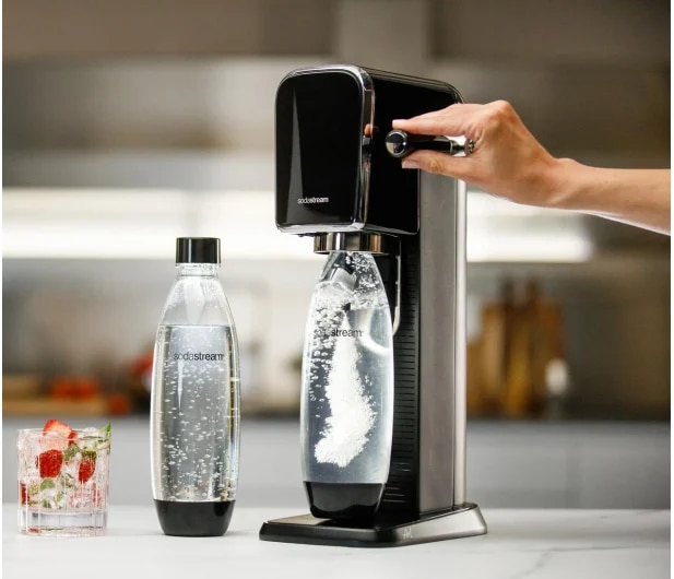Maszyna SodaStream z butelką, szklanką z truskawkami i ręką osoby ją obsługującej.