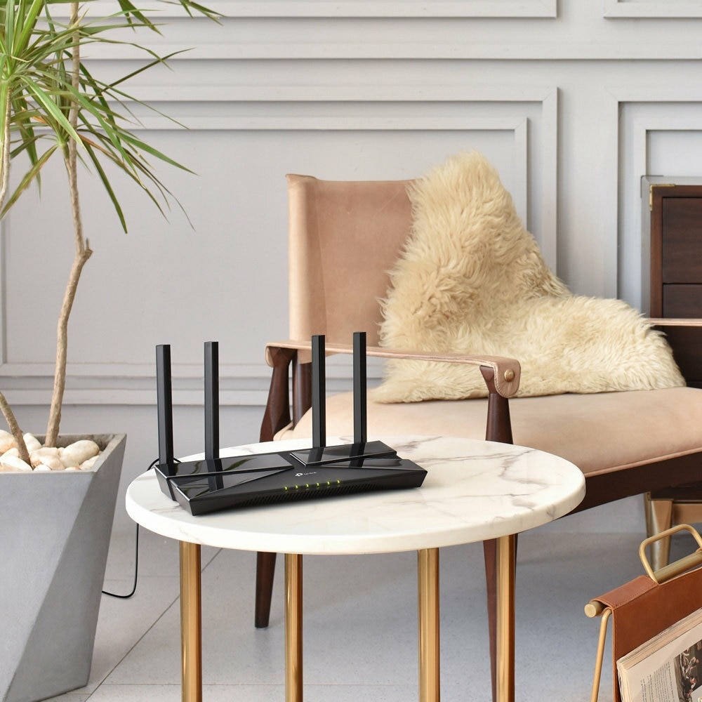 Czarny router z 4 antenami na marmurowym stoliku. W tle roślina doniczkowa i krzesło.