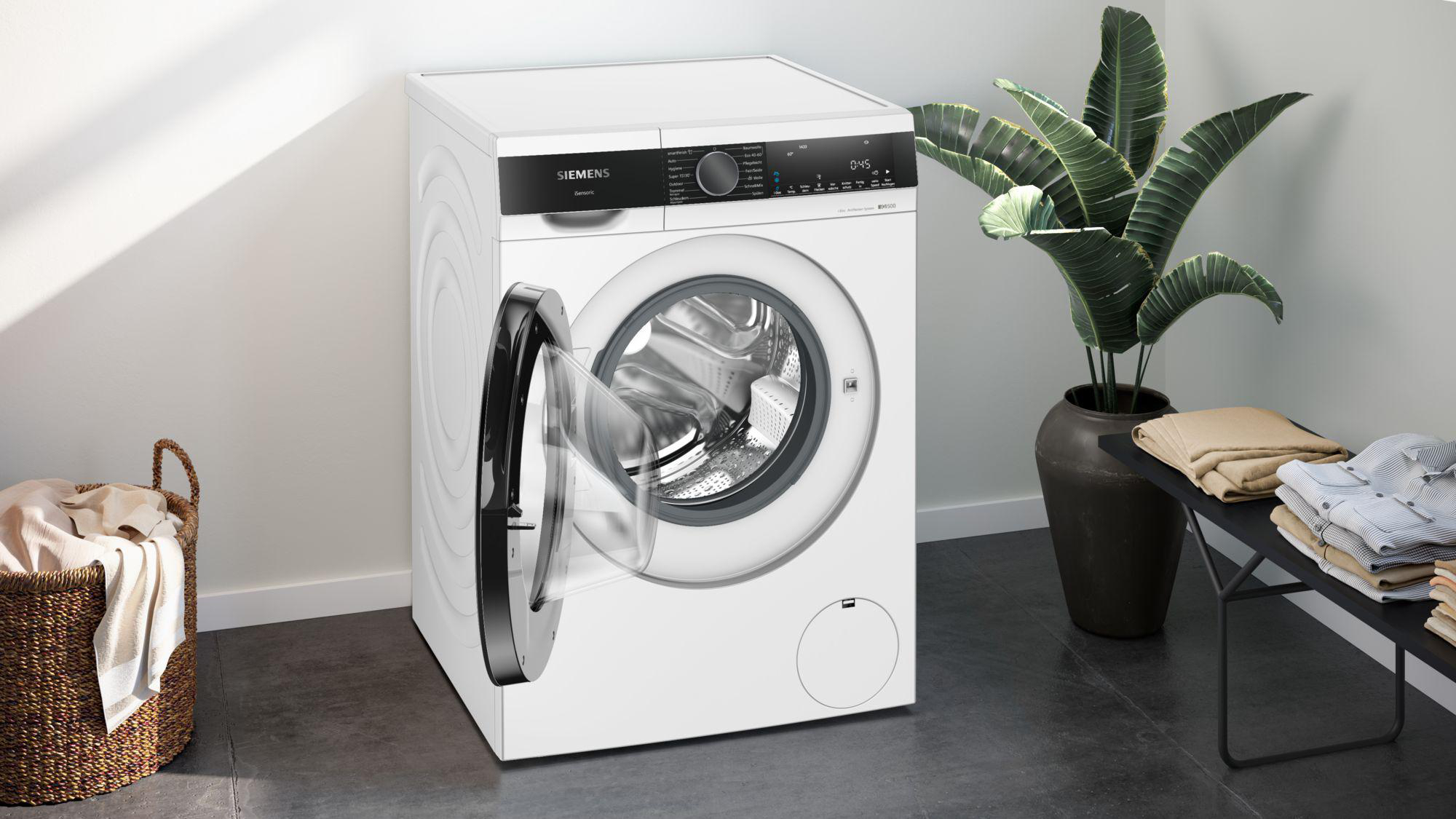 Eine weiße Waschmaschine mit offener Tür. Eine Pflanze in einem dunklen Topf, ein Wäschekorb und eine Bank mit Kleidung sind zu sehen.