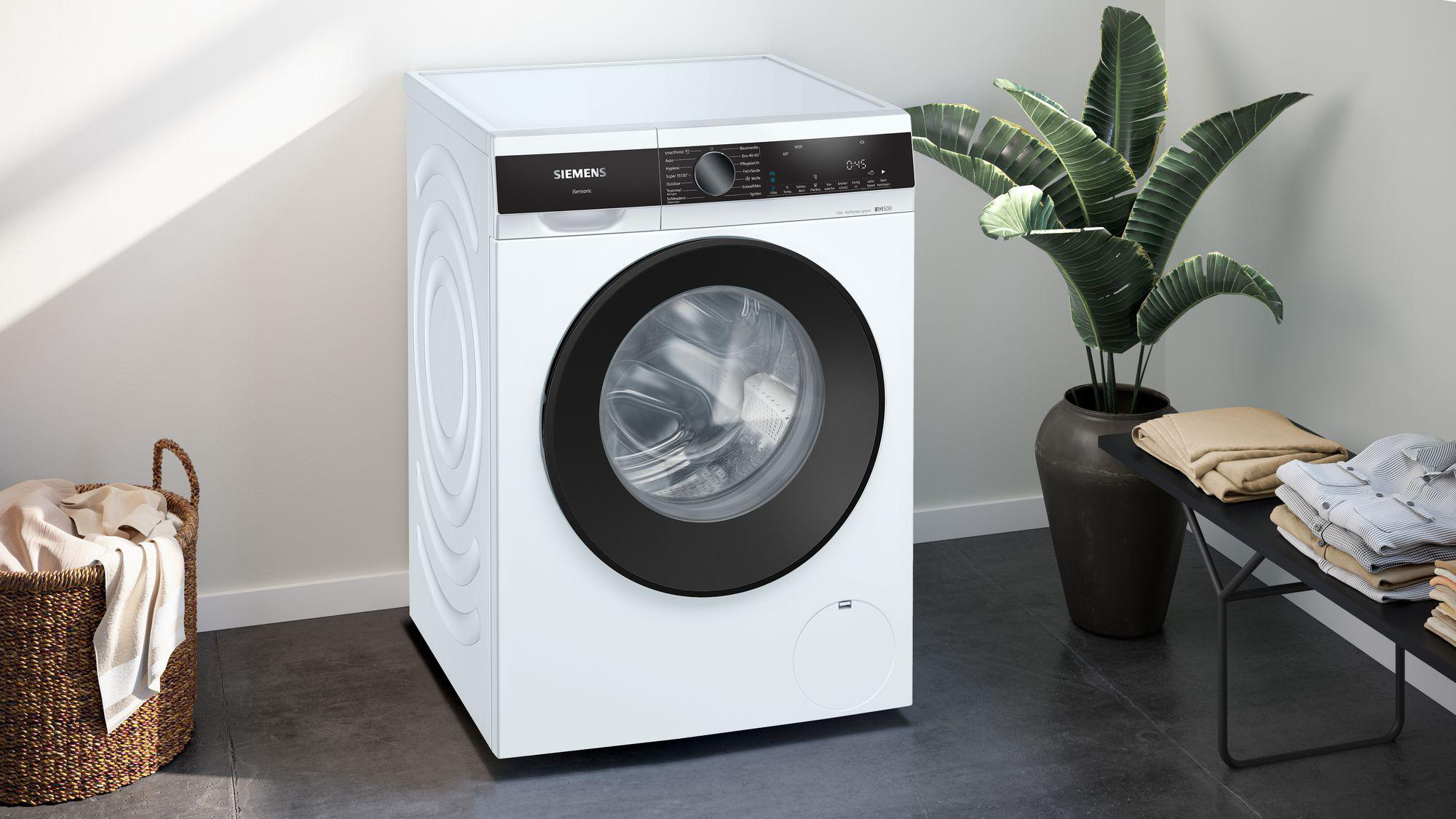 Eine weiße Siemens Waschmaschine mit schwarzer Tür, neben einer Pflanze im Topf und einem Korb mit Kleidung.