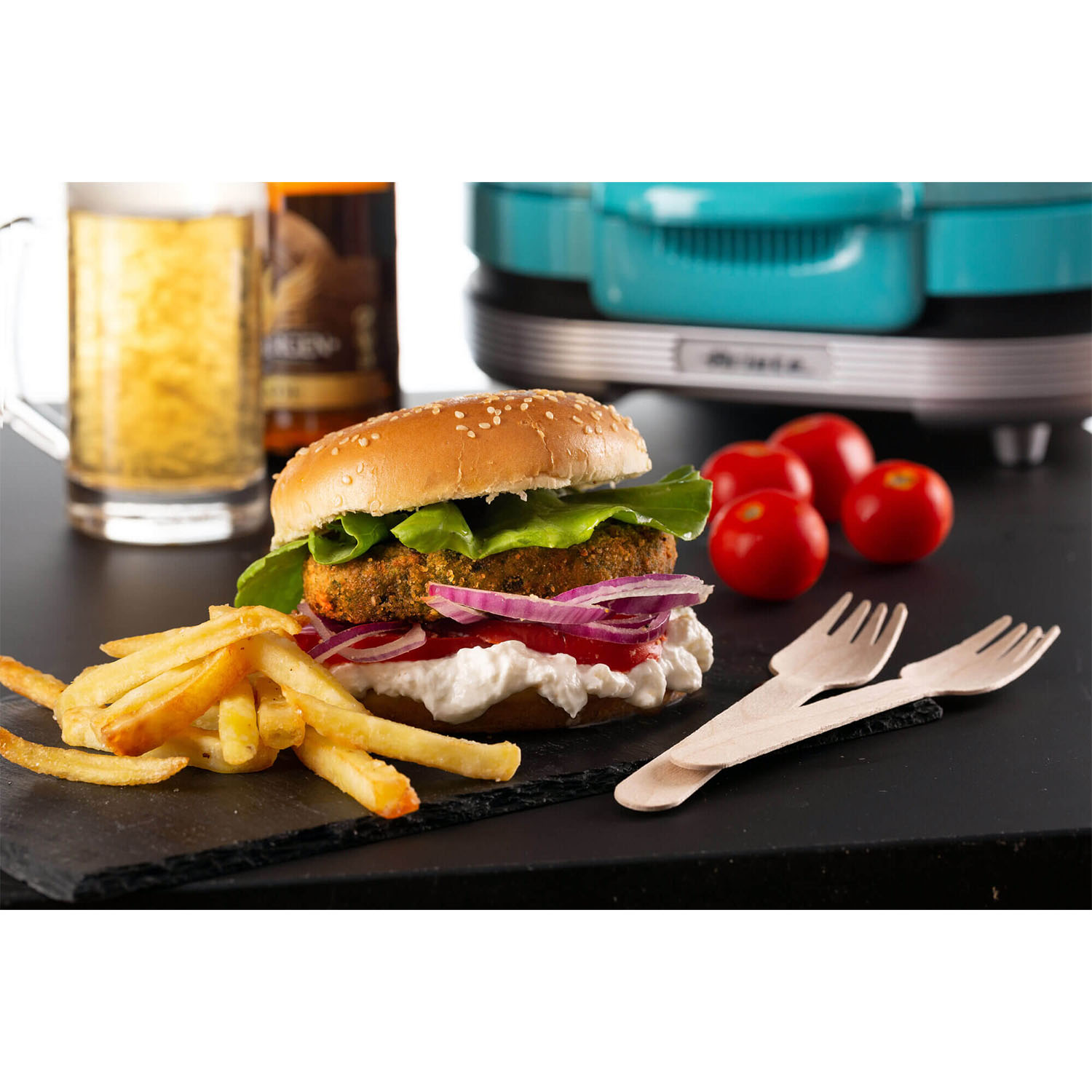 Un hamburger con patatine fritte, pomodori e birra su una superficie nera con un elettrodomestico verde acqua.