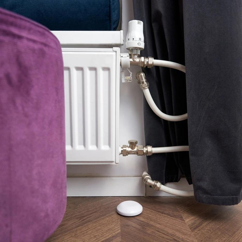 Fehér radiátor csövekkel és termosztáttal. Egy lila oszmán és fekete függöny van a keretben.
