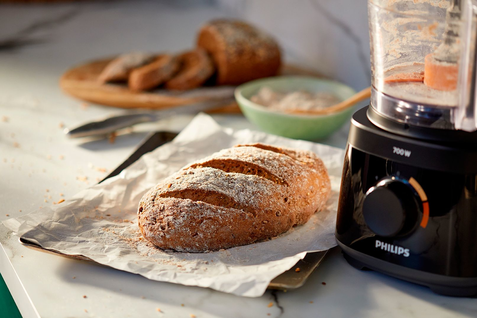 A loaf of bread on parchment paper with a food processor and sliced bread on a cutting board.