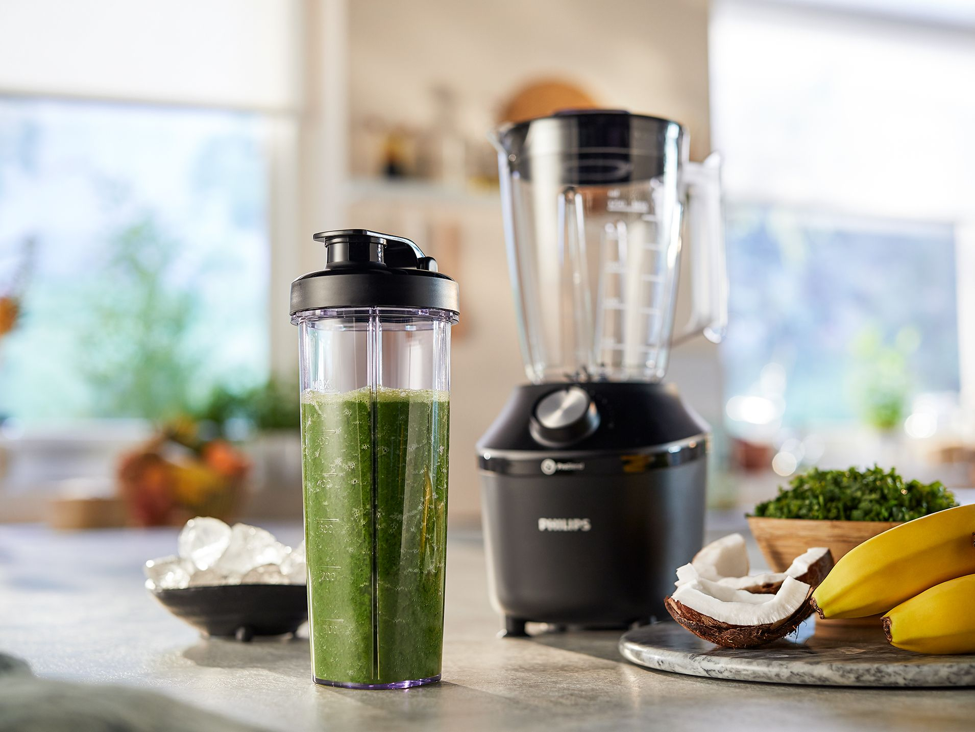 Blender with green smoothie, bowl of ice, coconut, and bananas on a counter.
