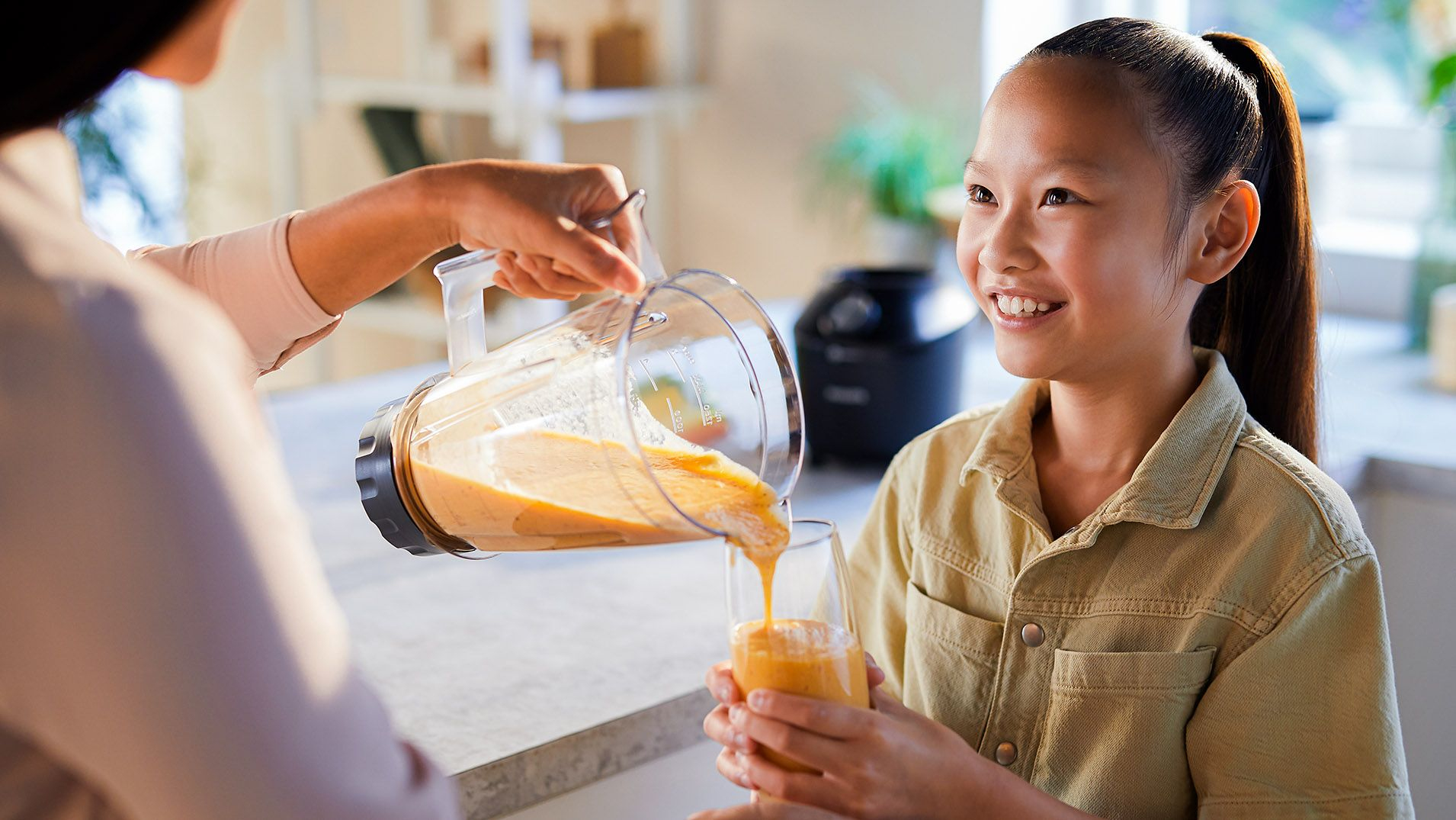 A girl smiling while someone pours juice into a glass.  A blender is visible in the background.