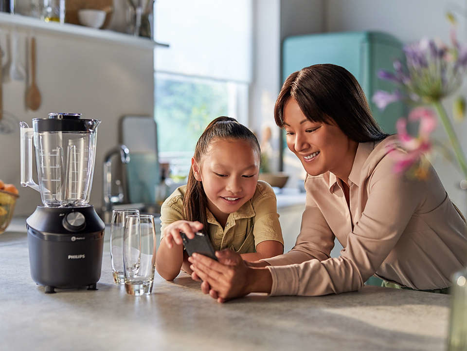 Woman and girl looking at a phone. Blender and glasses are in the kitchen.