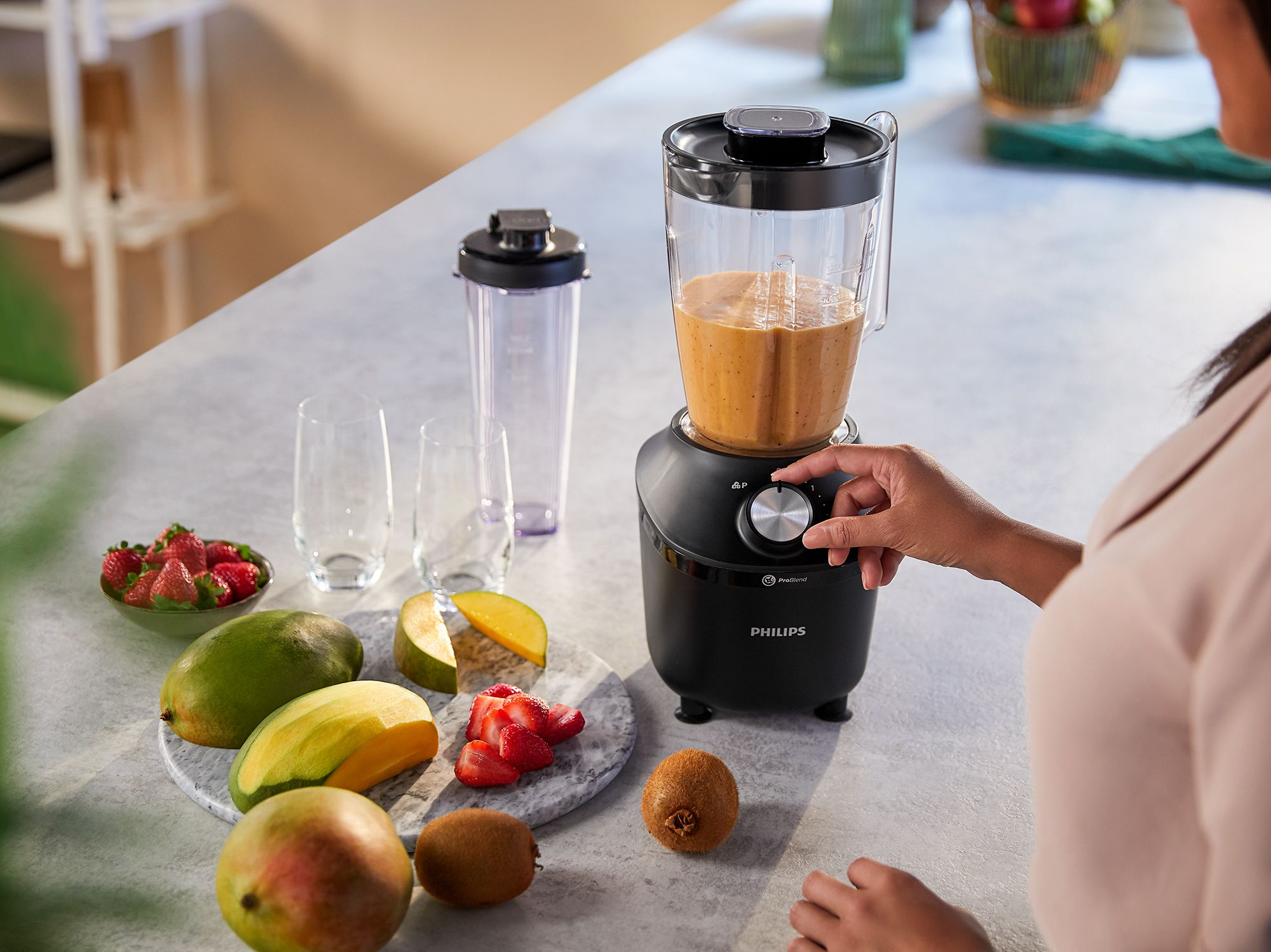 Blender with smoothie, fruit, and a person's hand adjusting the dial on a grey countertop.