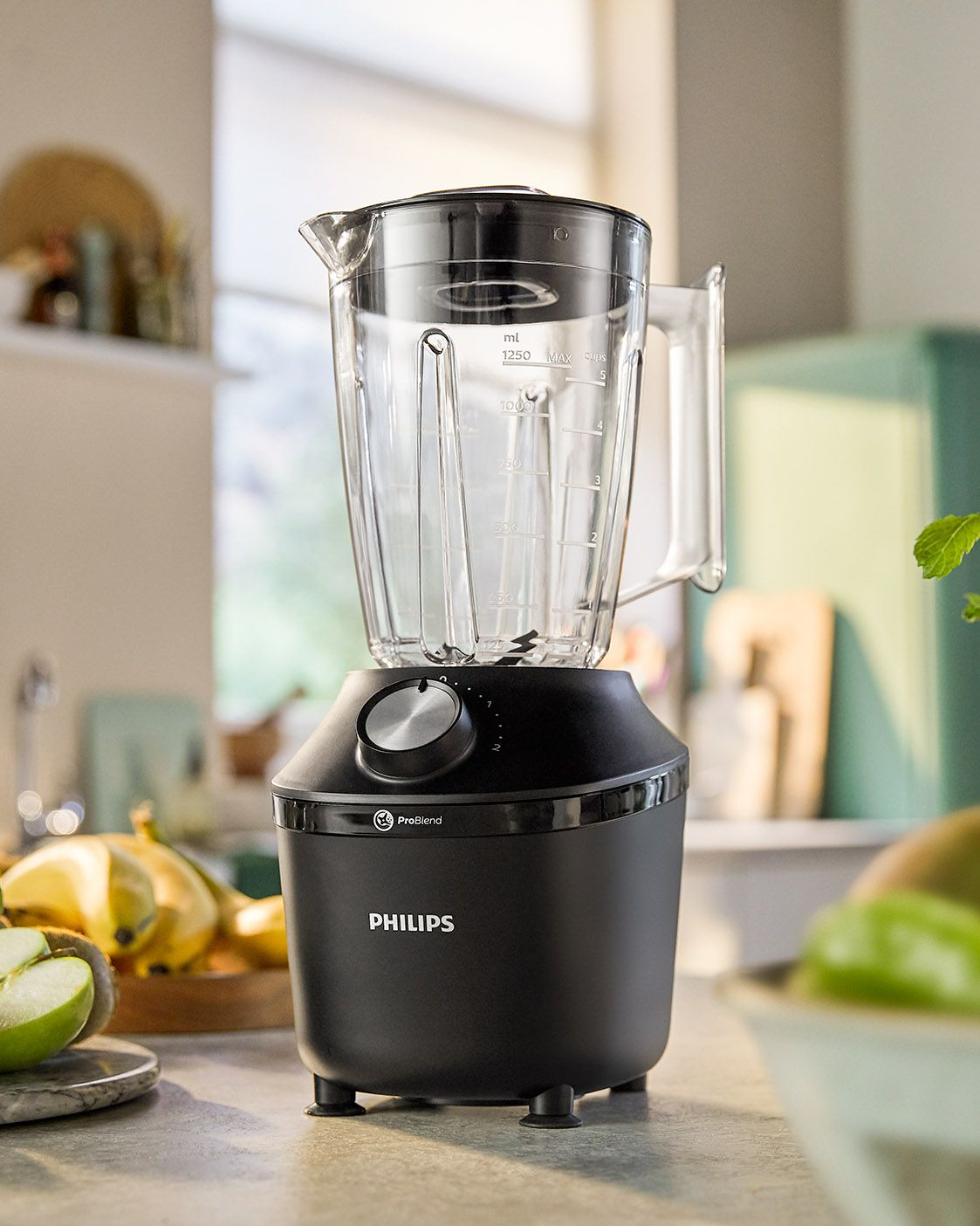A black blender with a clear pitcher. The blender sits on a countertop with fruits and a green wall in the background.