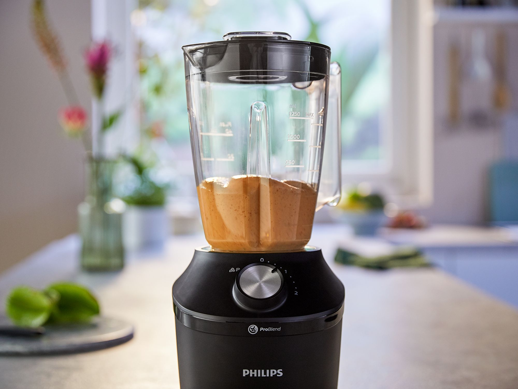A blender with a brown liquid inside. Black base with silver dial. Kitchen background.
