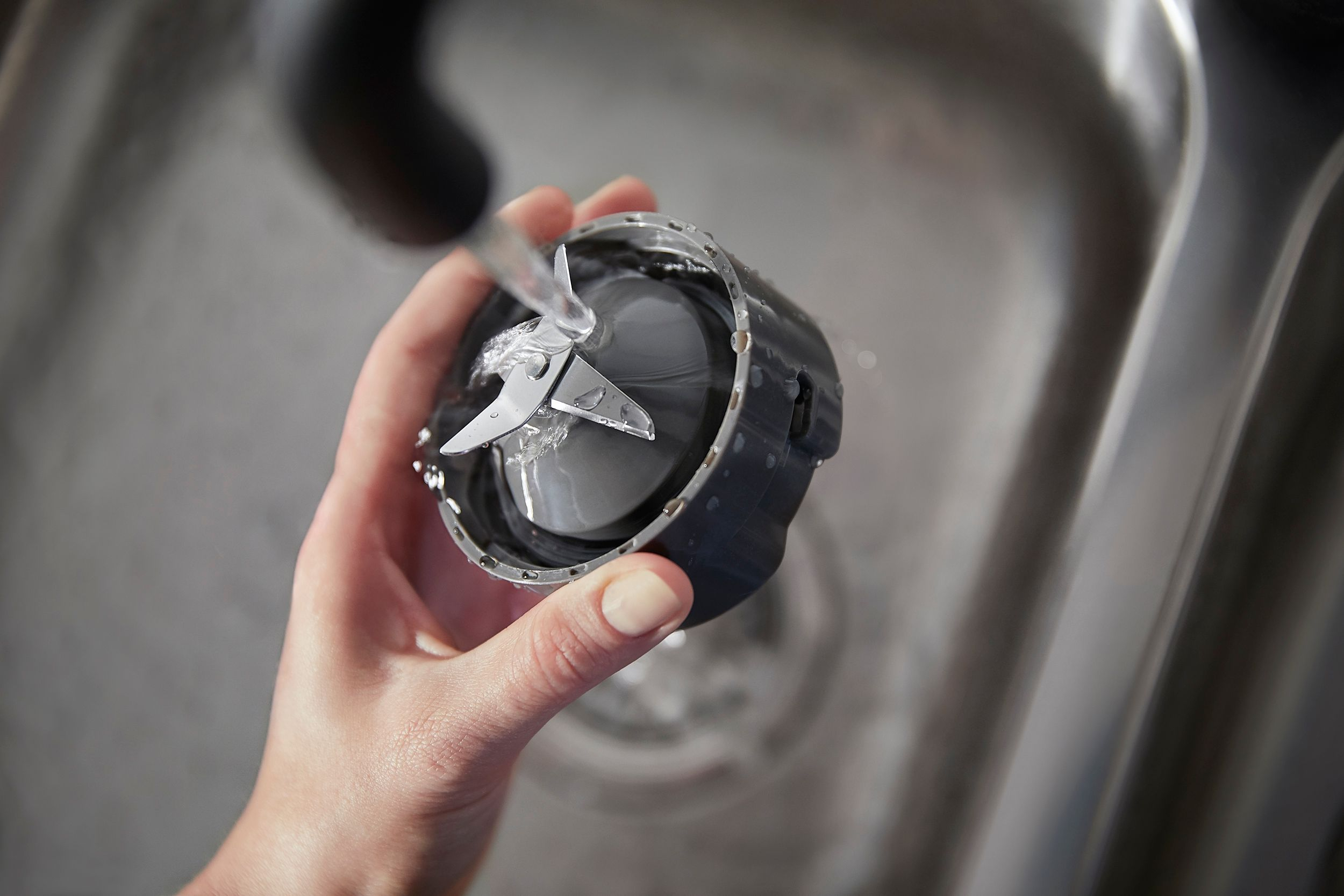 A hand rinsing a blender blade assembly under running water in a stainless steel sink.