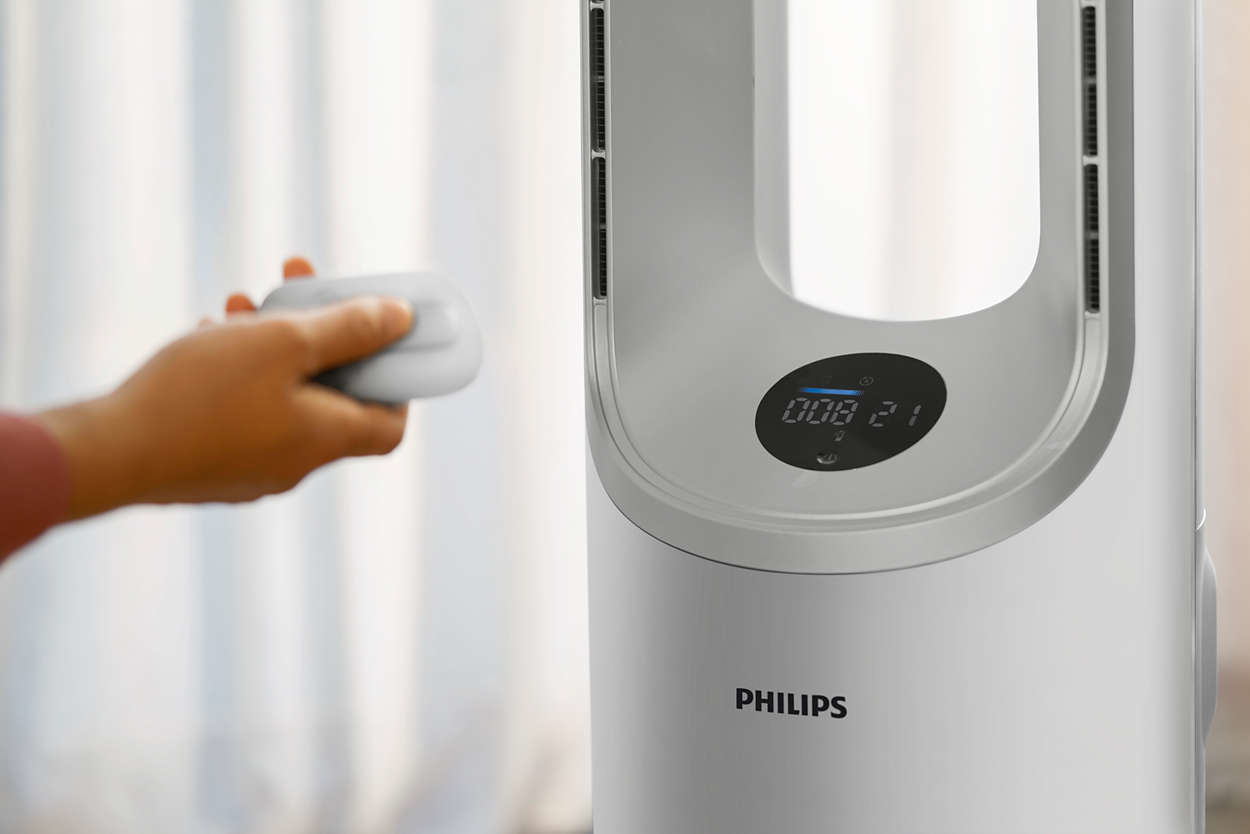 A hand holding a remote control near a white Philips fan with a digital display showing '008 21'.