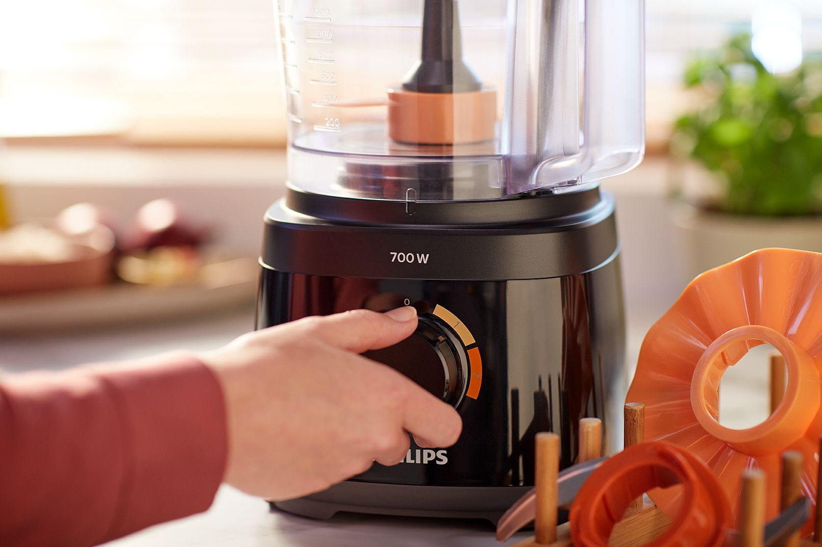 A person's hand turns the dial of a black food processor. Orange attachments are nearby.