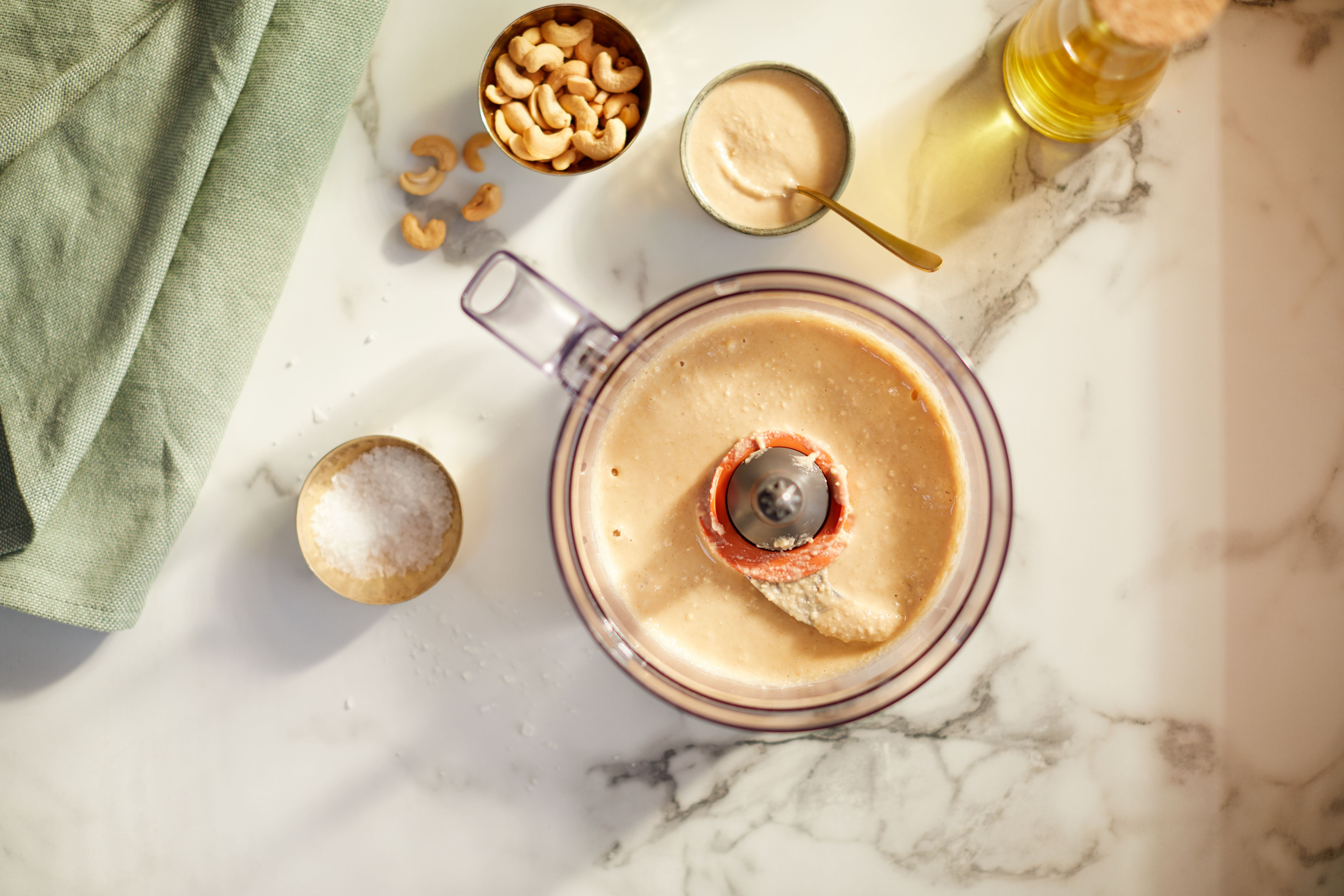 Overhead view of a food processor with cashew paste and other ingredients on a marble surface.