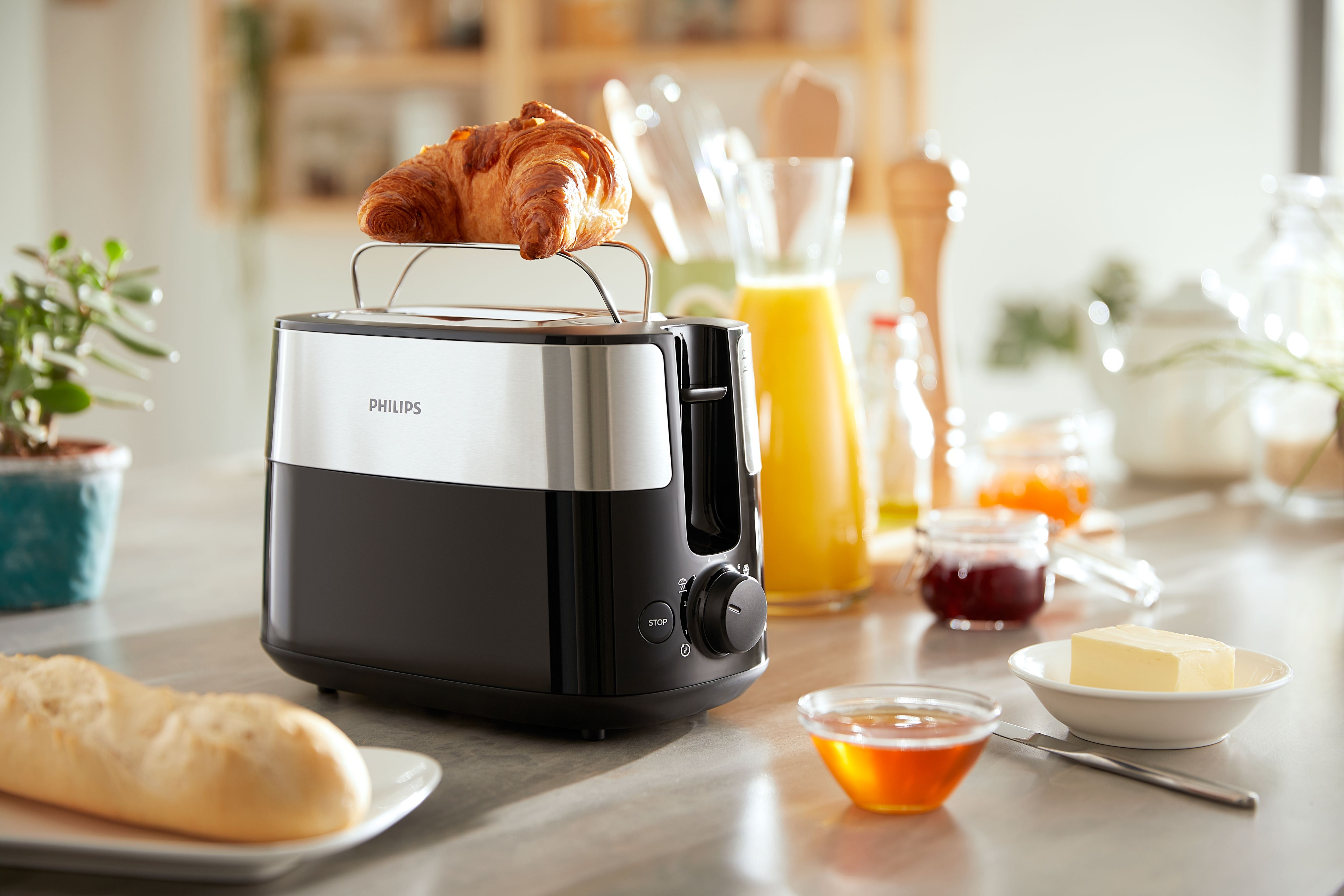 A toaster with a croissant on top, beside breakfast items on a countertop.