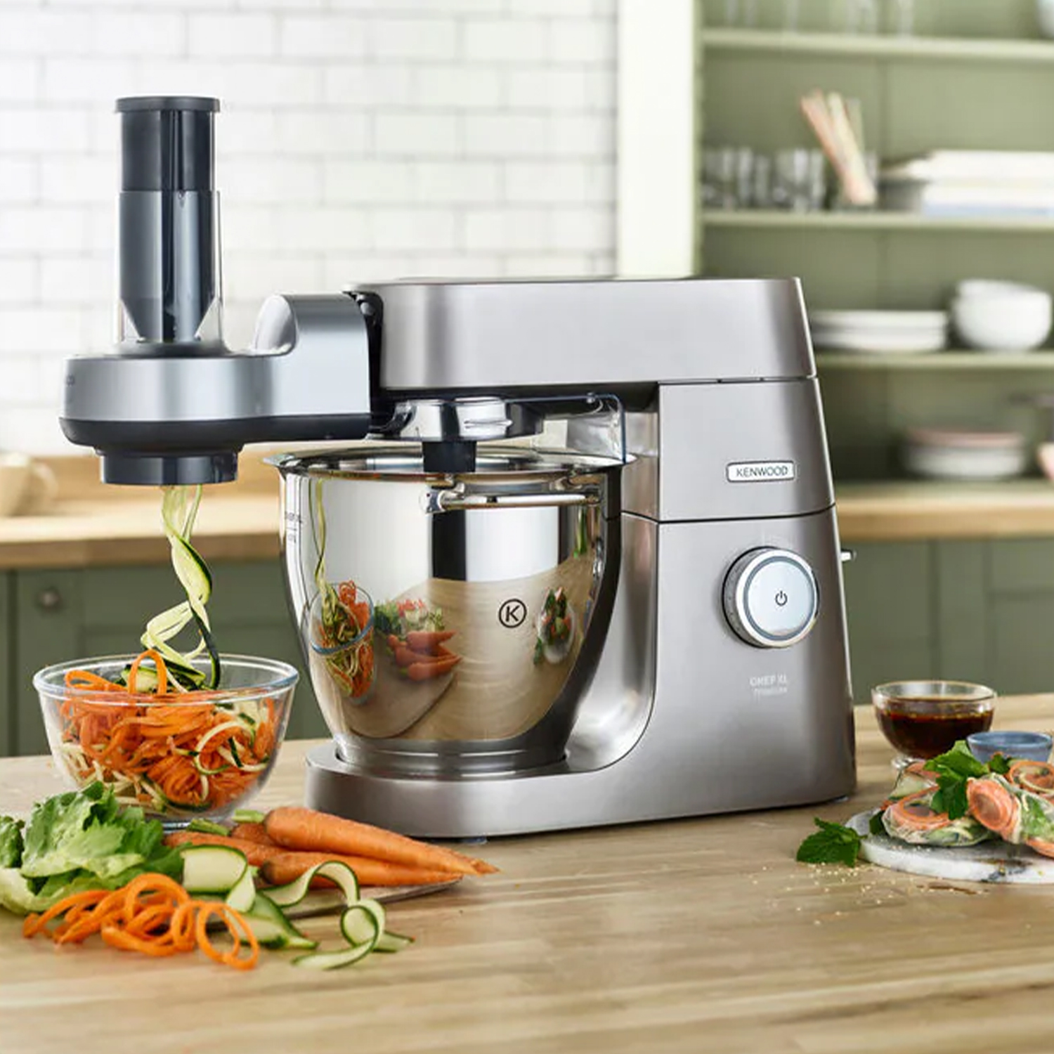 Un robot da cucina Kenwood argentato con cibo accanto su un tavolo di legno.