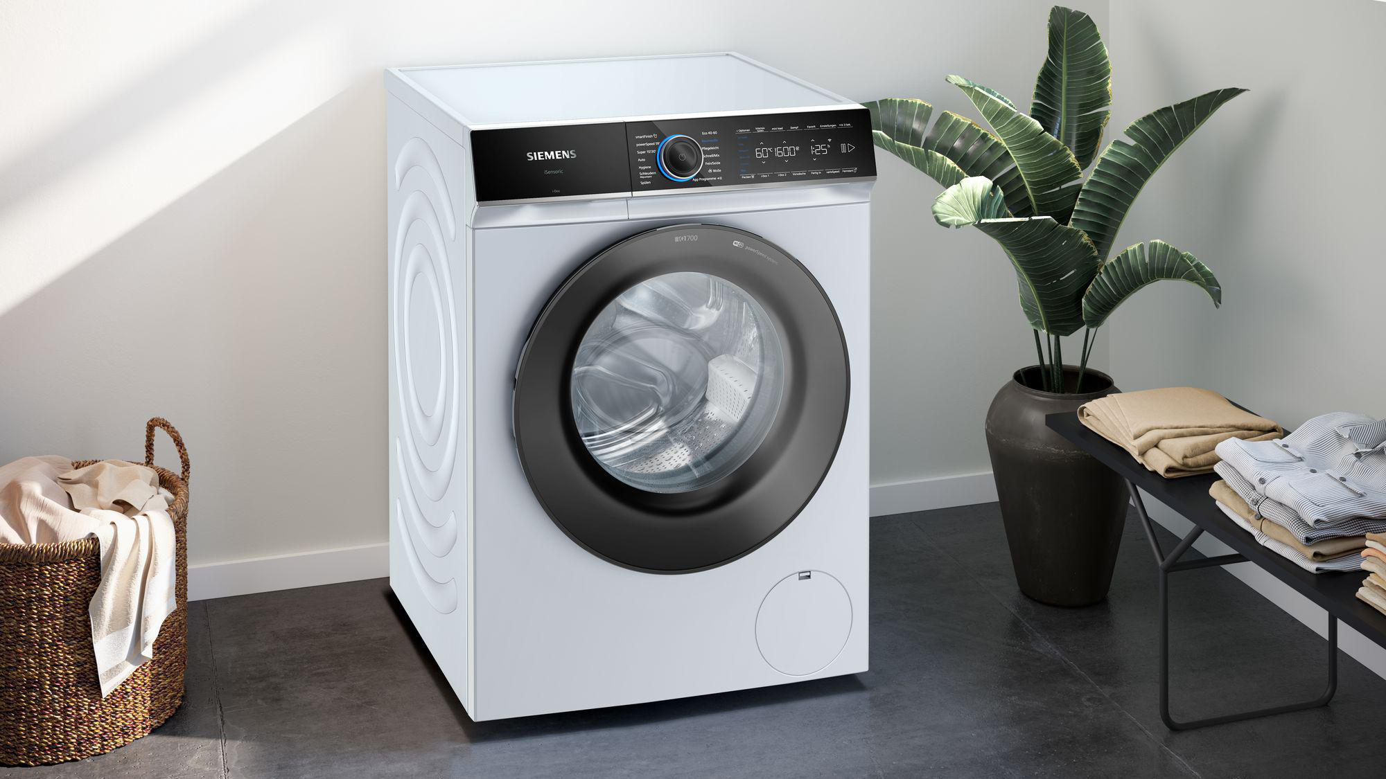 Eine weiße Siemens-Waschmaschine. Eine schwarze Tür und Bedienfeld. Daneben stehen eine Pflanze und eine Bank mit Kleidung.