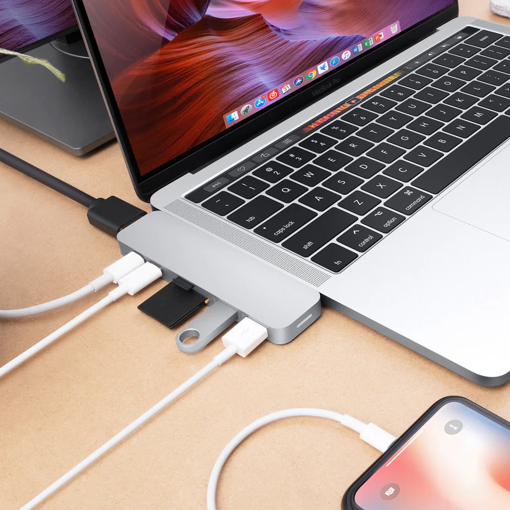 Portátil con un hub plateado conectado, con cables USB y un teléfono. Otro portátil en el fondo.